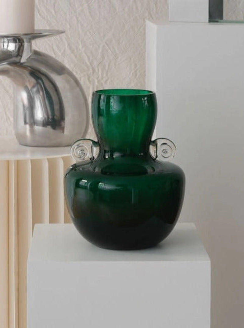 A Les Objets Amsterdam Vintage Bohemian Vase in dark green with two small clear glass handles on either side is displayed on a white pedestal. In mint condition, it stands against a backdrop featuring a shiny metallic decorative object and a fluted light-colored surface.