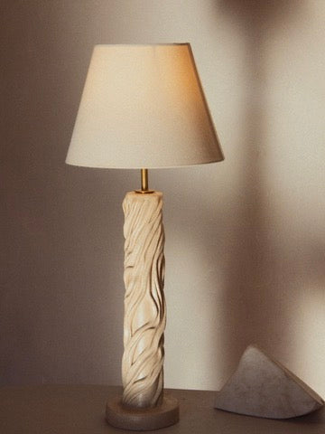 The "Waves" Table Lamp by Lorea Palacios, with its swirled ceramic base and beige shade, provides a cozy glow perfect for contemporary spaces. It stands on a flat surface, softly lighting the wall behind it. A small triangular white object is partially visible beside the lamp.