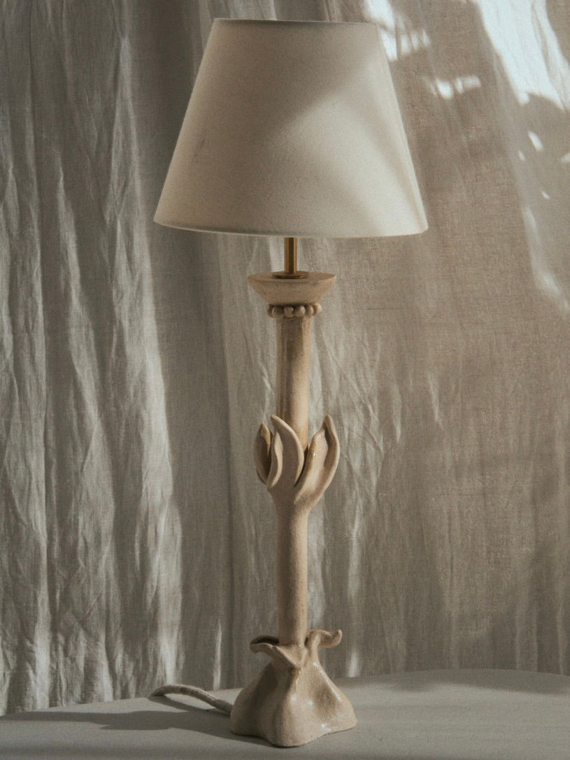 The "Bloom" Table Lamp by Lorea Palacios features a handcrafted design with a beige, plant-like textured stem and a matching beige lampshade, showcasing modern craftsmanship against a softly lit, sheer curtain backdrop.