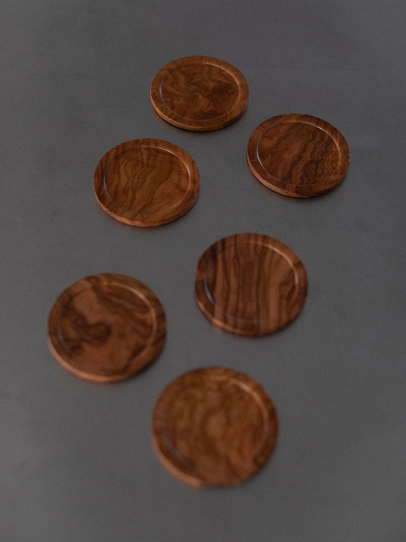 Six Out For Lunch Handcarved Coasters with visible grain patterns are arranged on a dark surface. The coasters exhibit intricate handcarved details and a smooth finish, showcasing natural wood tones from light to dark brown, offering a touch of natural elegance.