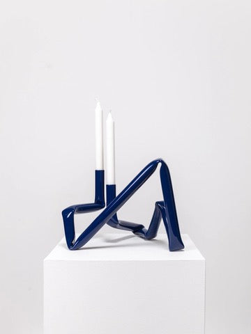 The Bucatini Candle Holder (Midnight blue) from AOT STUDIO, featuring an abstract winding design made from aluminum tubing, sits on a white pedestal. The handcrafted Denmark sculptural blue form elegantly holds two unlit white candles, standing tall against a plain white background.