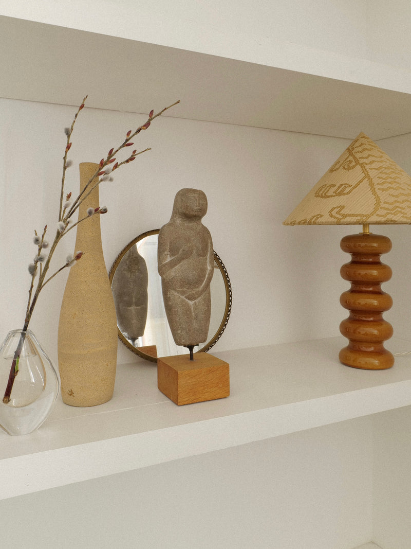A shelf displays a clear vase with branches, a beige ceramic bottle, the Marble Figurine Sculpture by Håkan Bonds from Collection apart, an Eva figurine, a round mirror, and a lamp with a stacked wooden base and a patterned beige shade.