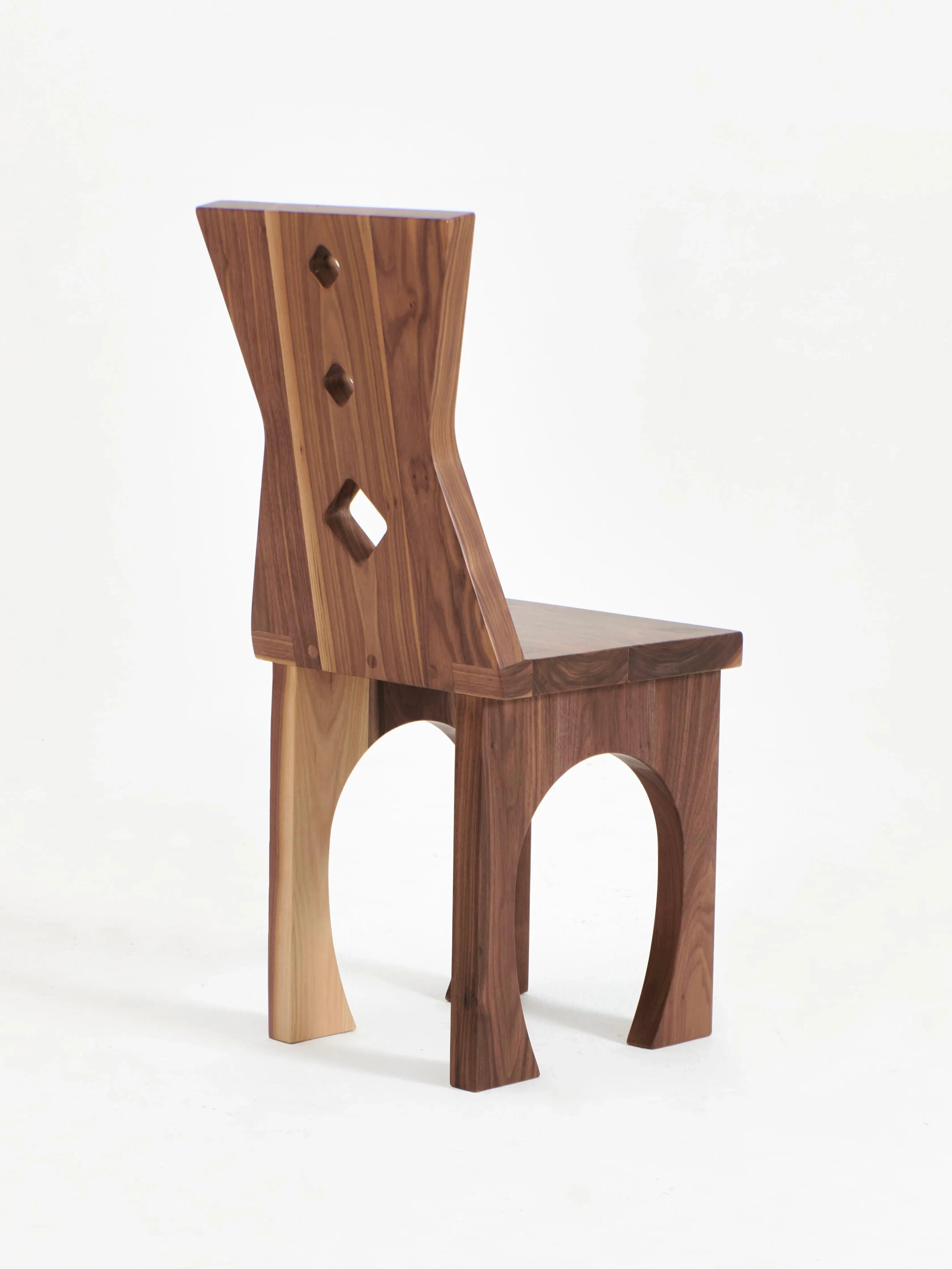 A wooden chair with a modern, geometric design, the Portugal Chair No 2 by Project 213A features an angular backrest with three diamond-shaped cutouts and a unique blend of light and solid walnut tones. The legs are arched, adding to the contemporary aesthetic. Handcrafted in Northern Portugal, it stands against a plain white background.