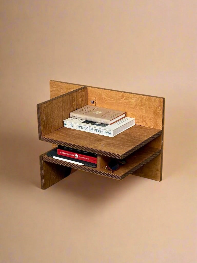 A wooden, wall-mounted shelf with a contemporary design features two levels that hold several books and a pair of glasses. Crafted from dark wood, this piece of functional furniture stands out against a plain beige background. (Inspired by Edoardo Lietti Studio products like the "Sisto" Versatile Wood Chair.)