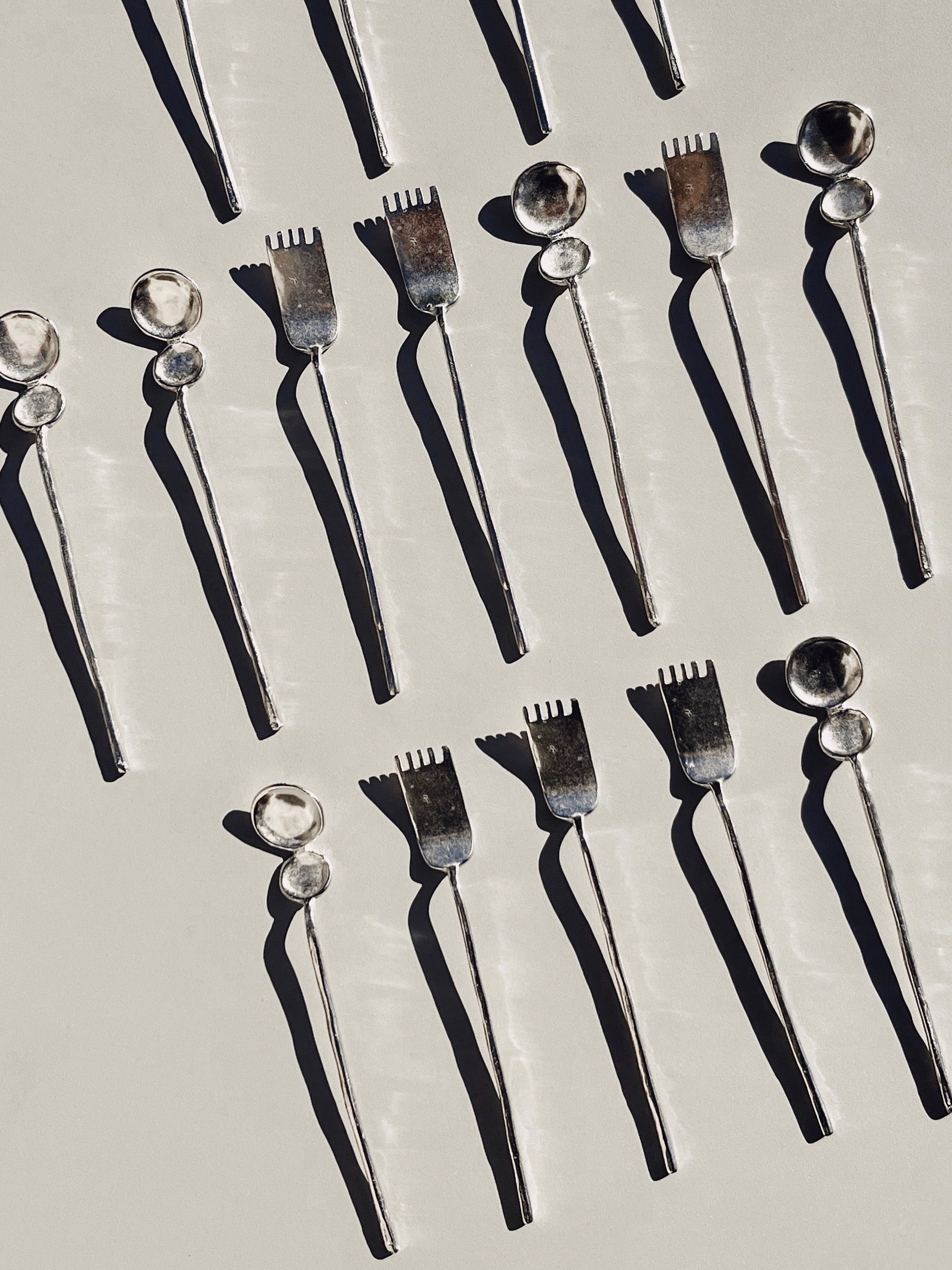 The "Artifact" Tea Cutlery - Duo by Hors-d'Oeuvre, skillfully handcrafted in France, is displayed on a light surface. This collection of spoons and forks features a neat, repetitive arrangement that forms an eye-catching geometric pattern. The shadows enhance the metallic texture of the utensils.