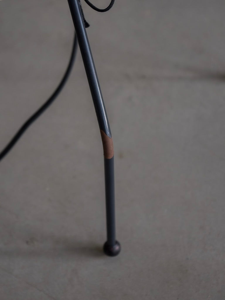 Close-up of a metal tripod leg on a gray floor evokes a mid-century modern vibe. A small brown patch appears near the base, wrapped with a black cable. In the unfocused concrete background, imagine an Isamu Noguchi - Akari UF by Bicci de' Medici completing the scene.