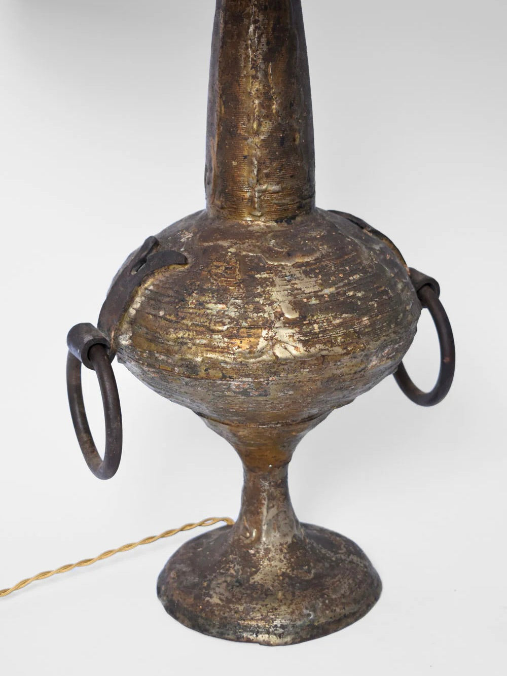 A rustic, antique Max Bre Gilded Iron Brutalist Table Lamp from the 1960s by Barracuda Interiors features a tall, narrow neck and a rounded base with two large metal rings on its sides. The surface is weathered with a patina finish, and an electrical cord is visible at the bottom.