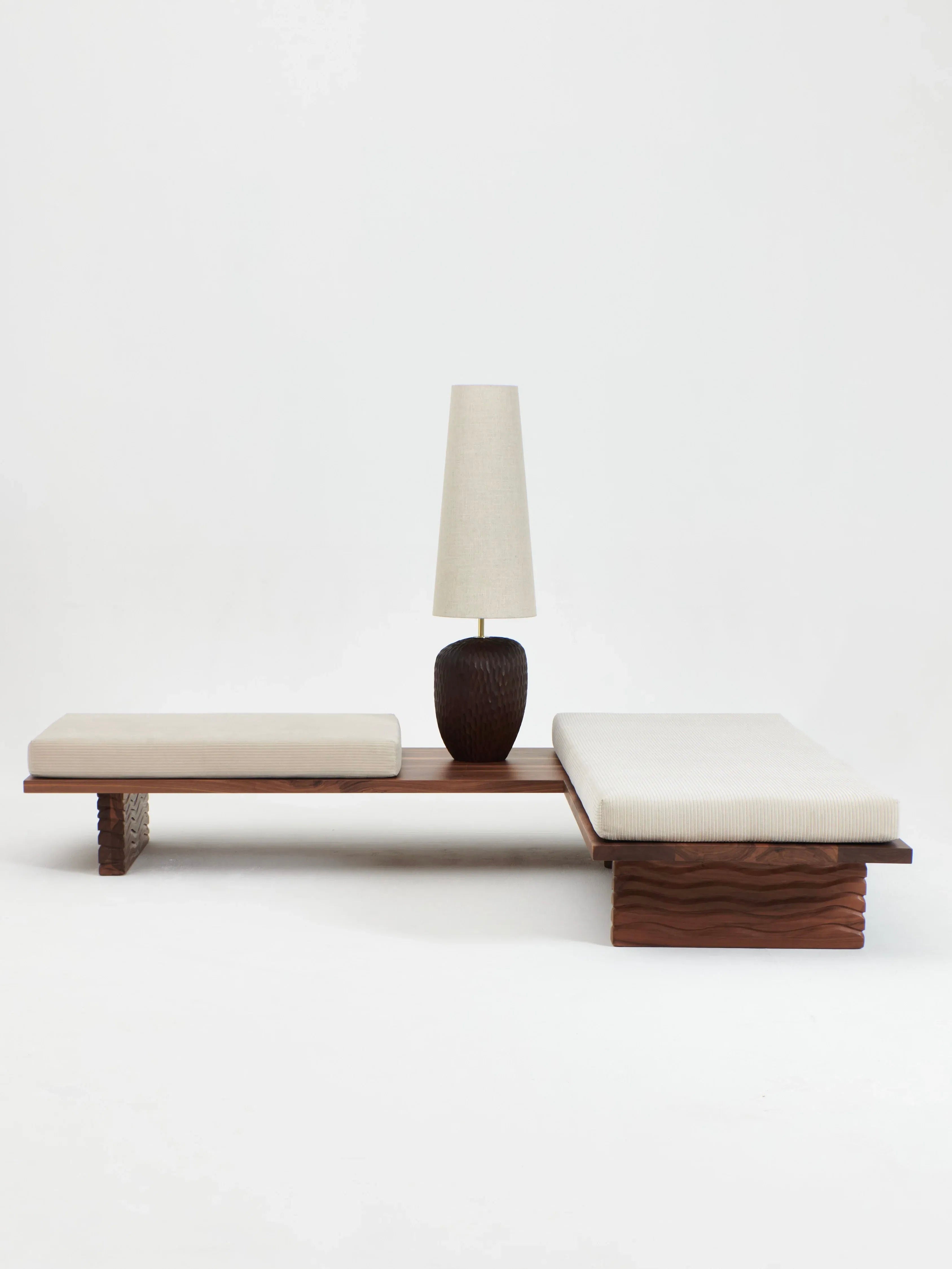 A minimalist furniture setup is defined by the Nazaré Daybed L Shape from Project 213A, a low platform expertly crafted from solid walnut. The daybed features two removable beige cushions and is complemented by a tall table lamp with a dark base and light shade. The elegant scene is set against an off-white backdrop, showcasing pieces that are beautifully handmade in Portugal.