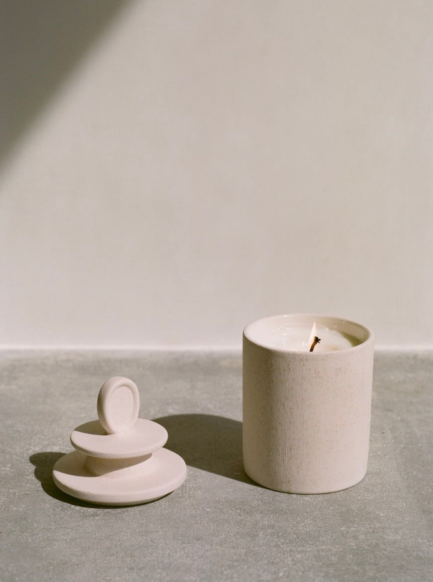 A Astrology Sign Candle by Olfactif in a textured holder next to a stack of small handmade ceramics, all placed under soft sunlight on a smooth surface.