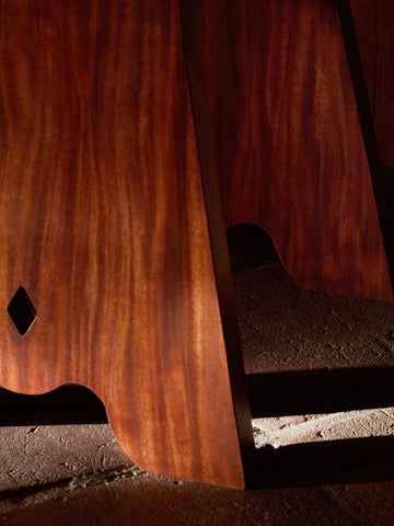 Close-up of the Rambling Chair in rich African Mahogany by Lemon, featuring elegant curves and a natural grain pattern reminiscent of a classic milk stool. Sunlight casts a soft shadow on the floor beneath, embodying contemporary elegance.