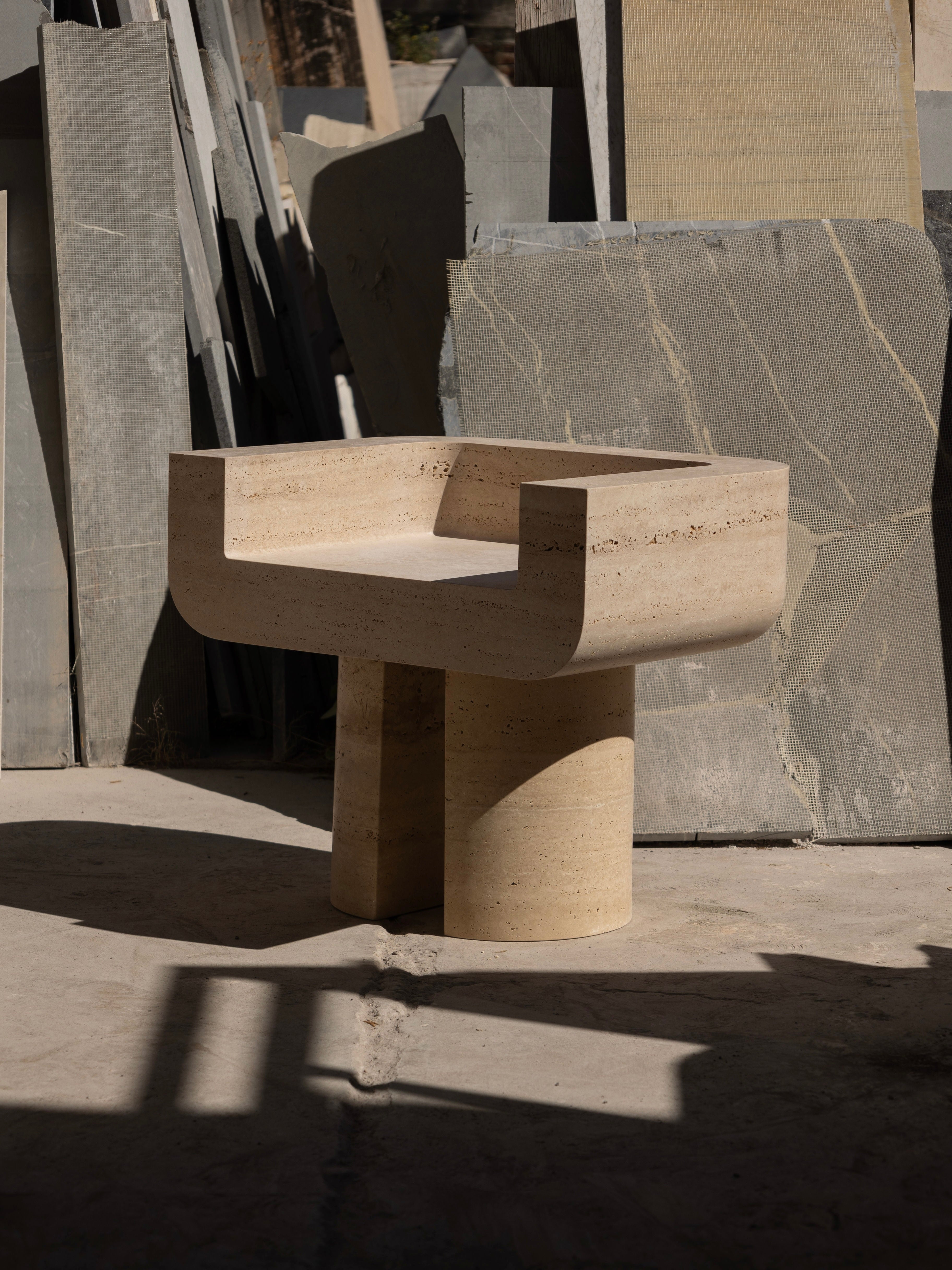 The M_001 Chair by Monolith features a modern, minimalist design with a stone-carved square seat and curved armrests on a concrete floor. Its geometric gray and beige vertical slabs create intriguing shadows, enhancing the chair's versatility as indoor-outdoor furniture.