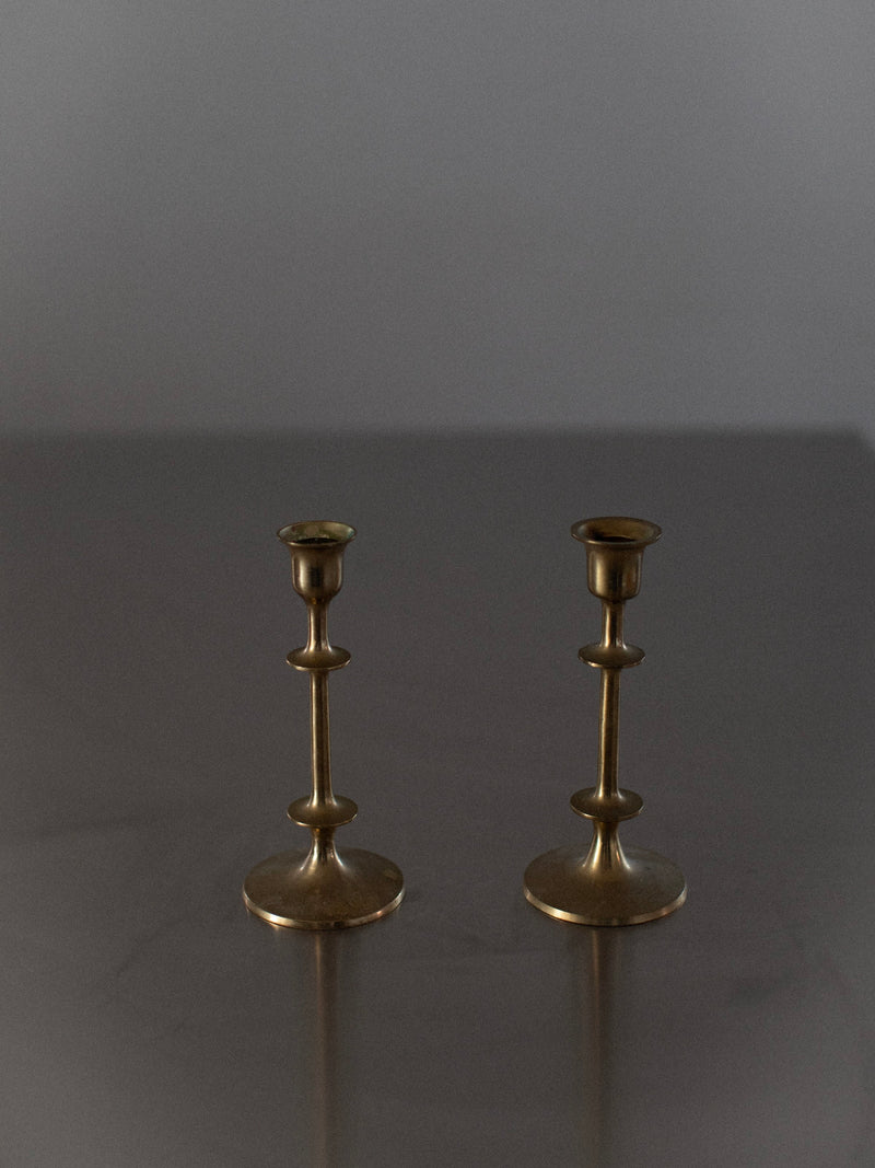 Two Brass Candleholders by Out For Lunch stand on a reflective gray surface. The candleholders, featuring a simple yet elegant design with rounded bases and tiered, slender central columns, capture the essence of both classical and contemporary design. The background is plain and softly lit.