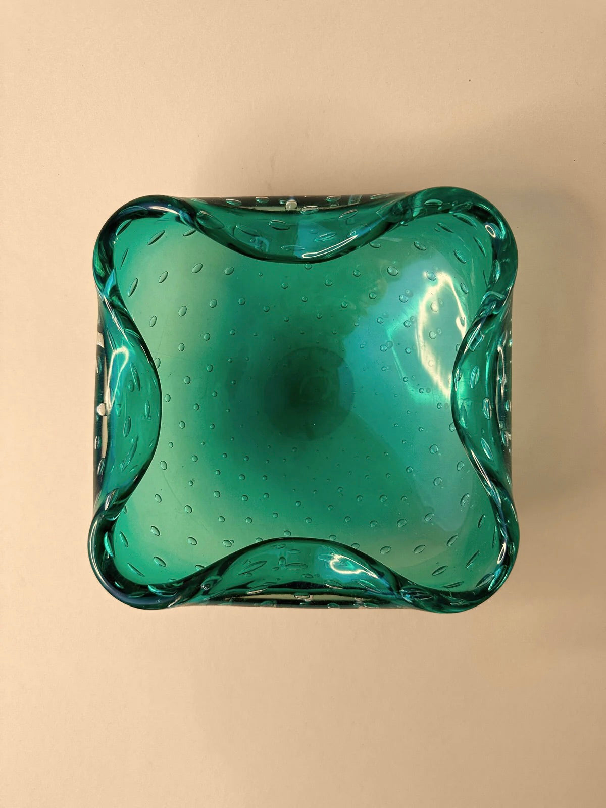 A top-down view of a mint green Murano ashtray from the 1960s, featuring an undulating, square shape. This Dodo Vintage piece showcases craftsmanship reminiscent of Barovier Toso, with a textured pattern adorned by small, raised bullicante bubbles scattered across its surface. The background is a plain, light beige color.