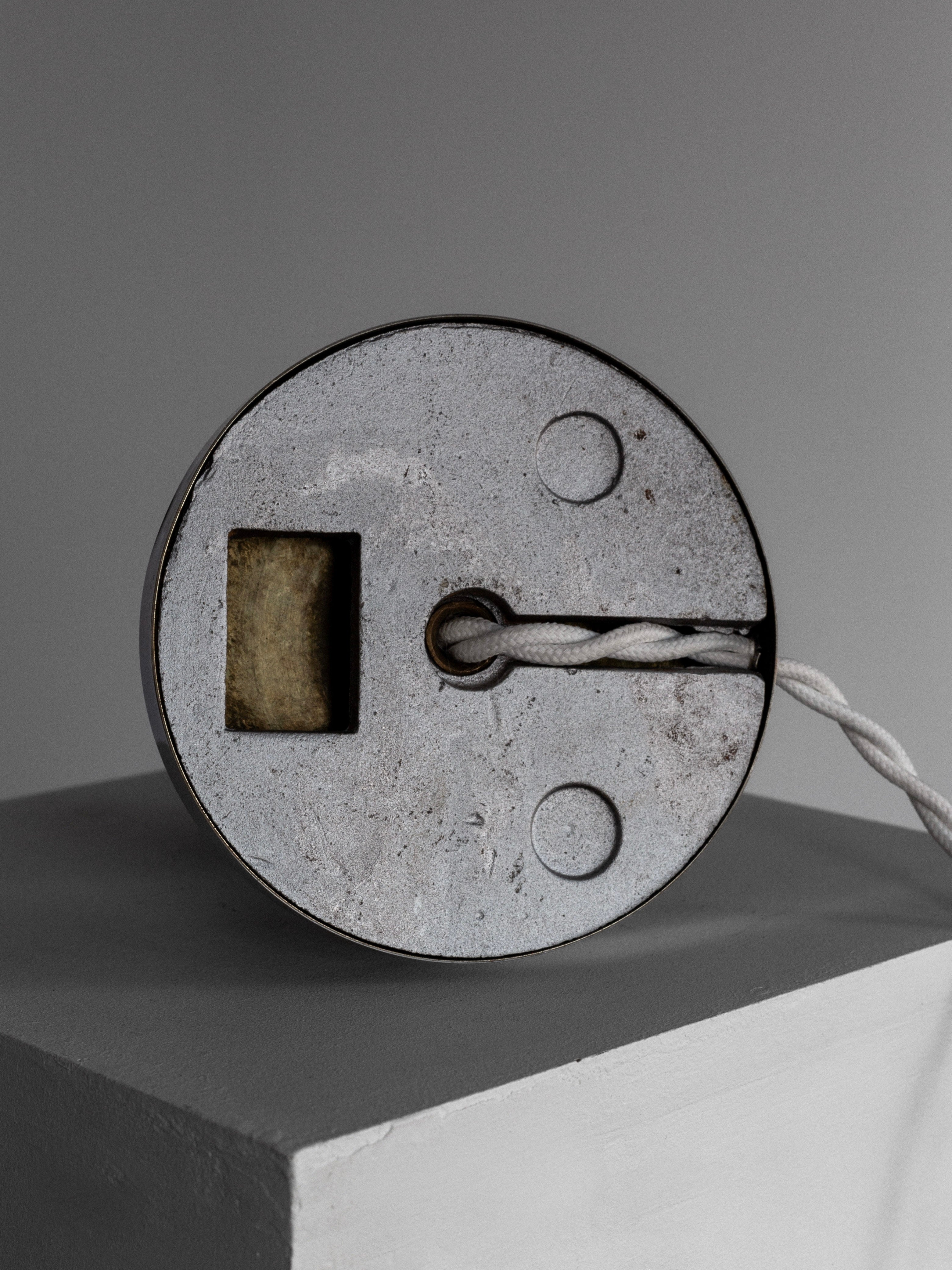 A close-up of the Wood & Chrome Table Lamp, 1930s by Spigel captures the sleek elegance of 1930s Sweden with its round metal design featuring a circular groove. A white cord threads through its center, reminiscent of an Art Deco style. Resting on a gray and white geometric surface, the softly lit background enhances its chrome allure.