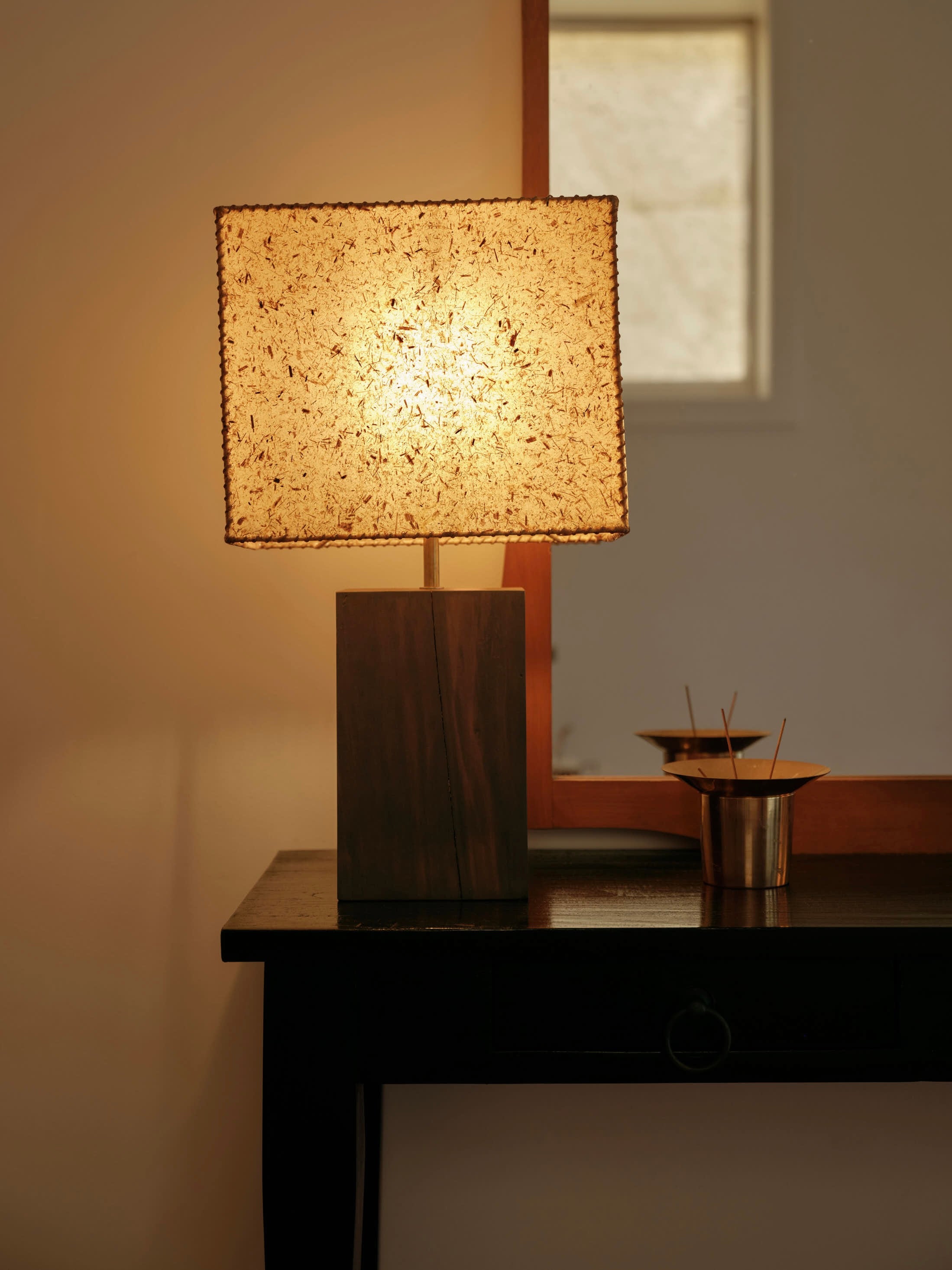 A warmly lit Lana Launay 1948 Table Lamp with a textured beeswax shade stands on a dark wooden table. Behind it is a mirror. To the right, there's a metallic container holding sticks for aroma diffusion, adding a cozy ambiance to the setting.