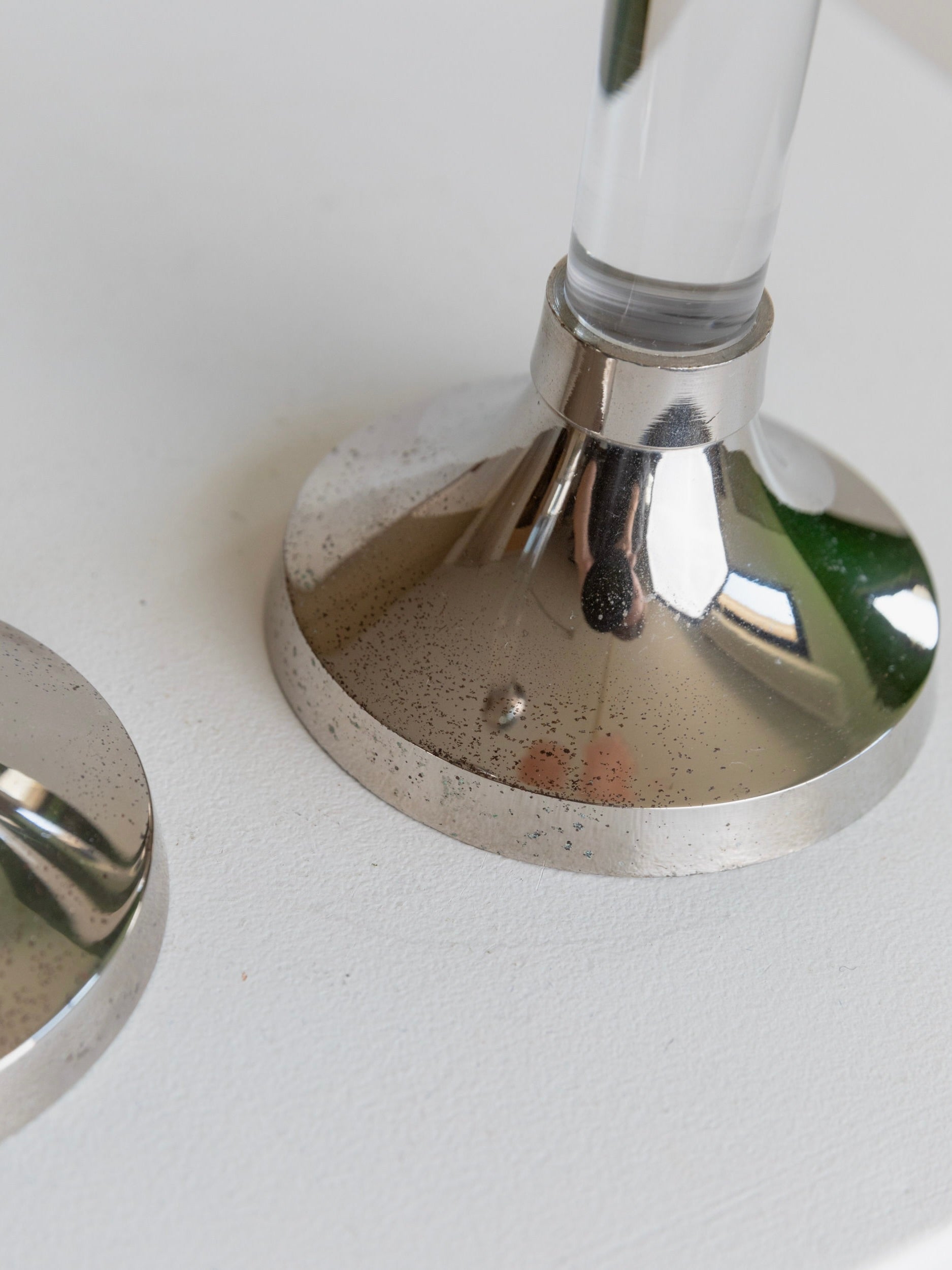 Close-up of two chrome-plated metal bases from the Bottega Jacobs Set of 2 Candleholders on a white surface. One base supports a transparent acrylic glass rod, creating an elegant contrast, while the other remains empty. The metallic surfaces feature a speckled texture and subtle light reflections, reminiscent of vintage candlesticks.