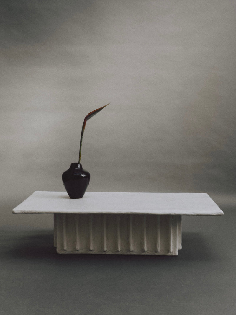 Experience minimalism with the Columnar Coffee Table by Ombia Studio: a black vase with a single long-stemmed leaf on a white, textured rectangular pedestal against a muted gradient background. Available in custom sizes and made to order for your space.