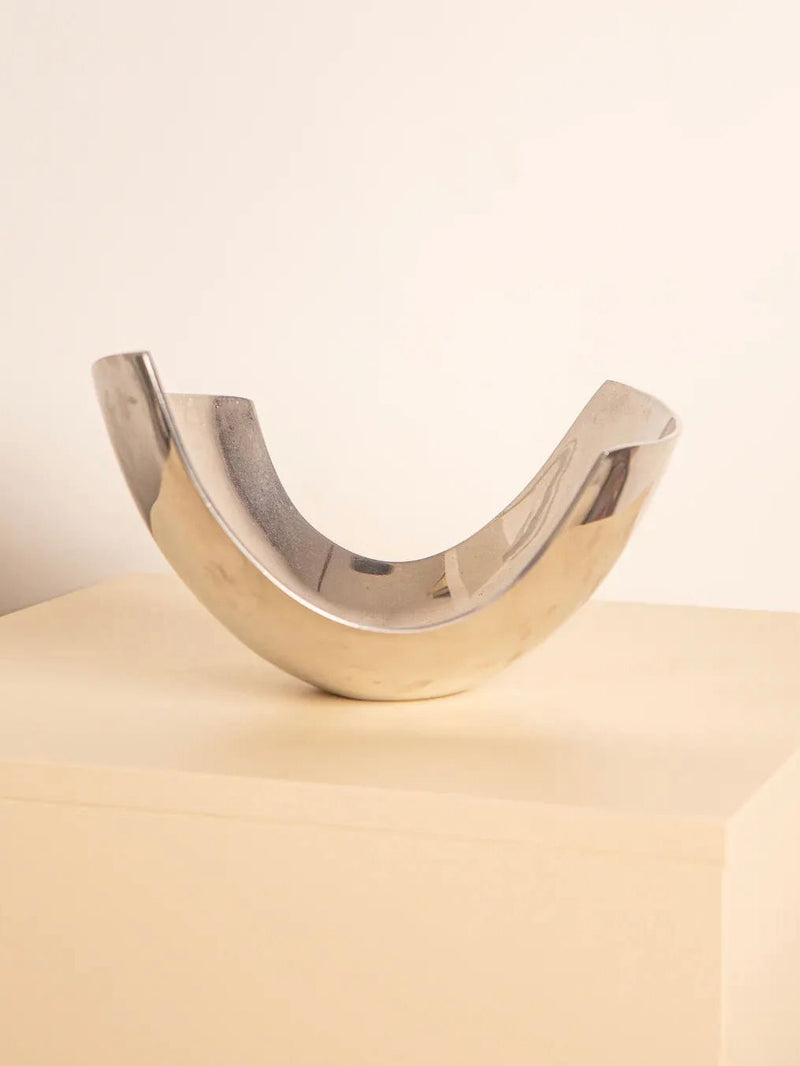 The "U"-Shaped Fruit Basket 80's by Treaptyque, featuring a retro design with a reflective curved metal structure, sits on a light wooden surface against a simple beige background. This sculpture-like piece boasts smooth and shiny surfaces that form a semi-circular shape.
