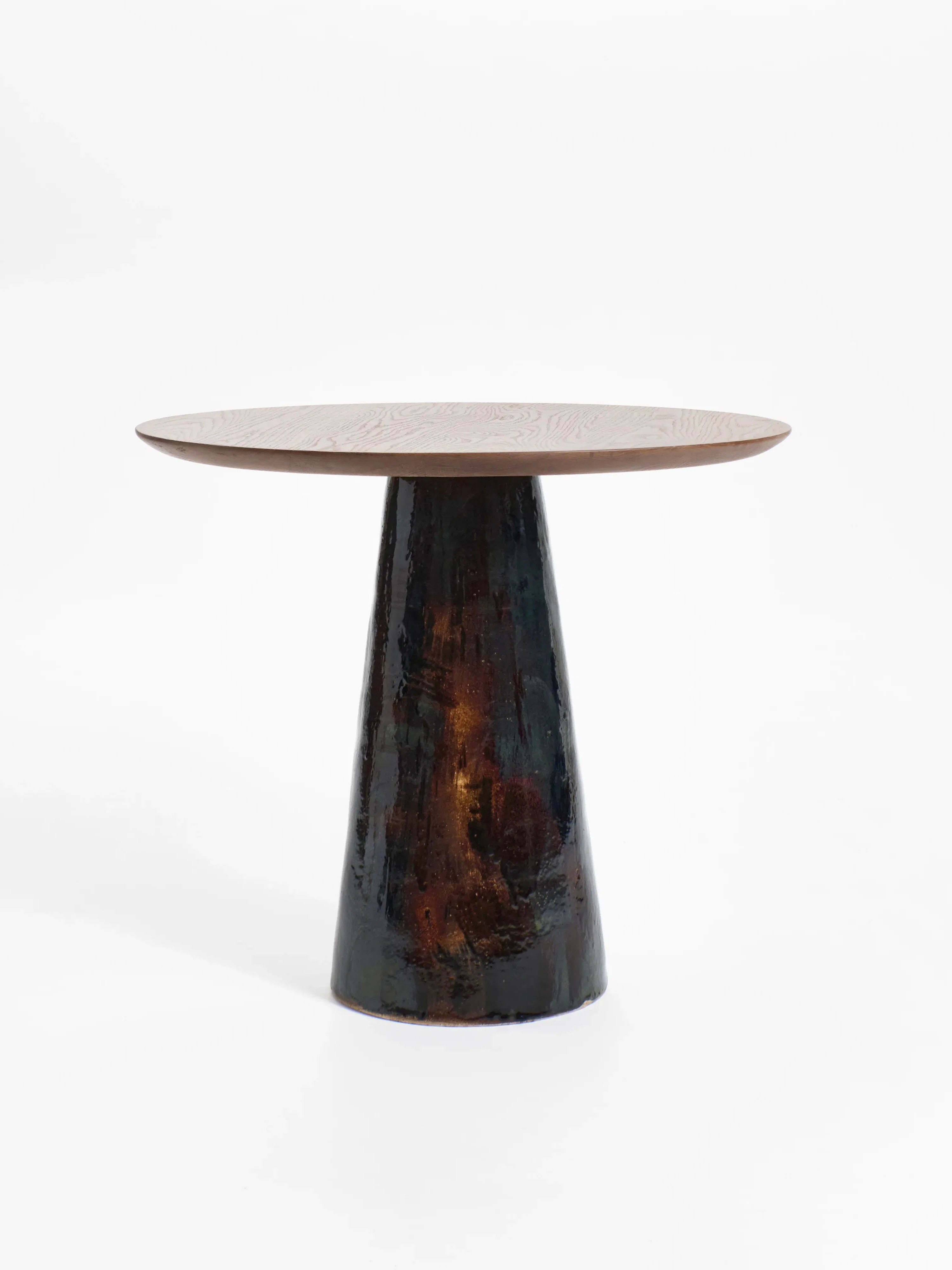 The Porto Belo Table by Project 213A features a small round wooden top and a striking hand-sculpted ceramic base, ideal for intimate bistro dining. Set against a plain white background, it exudes elegance through simplicity and craftsmanship.