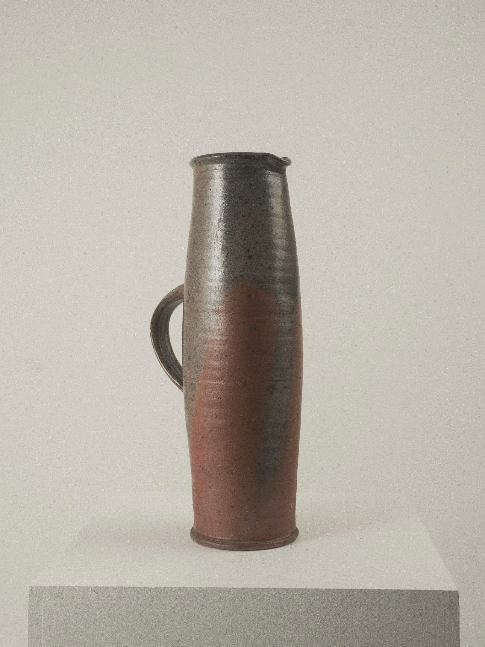 The Large Pitcher by Gustave Tiffoche from Figuresdesign is a tall, cylindrical ceramic jug with a handle and features a dark gray to reddish-brown gradient. Displayed on a white pedestal against a light gray backdrop, it radiates timeless French elegance.