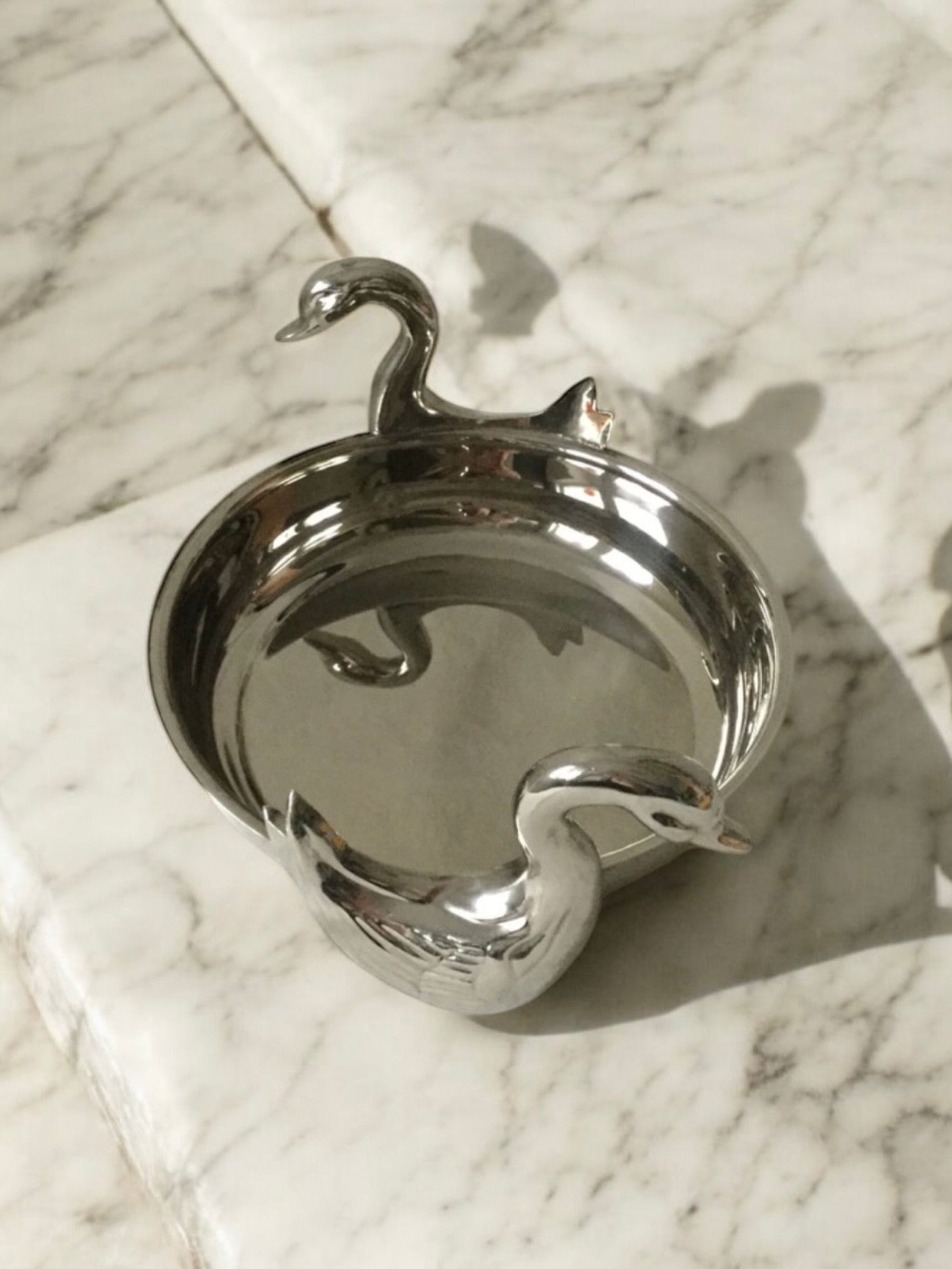 The Twin Swan Dish by Flock Studio, with silver plating and sculpted swan heads on its rim, casts a shadow on the marble surface in soft natural light, ideal for serving sweets elegantly.