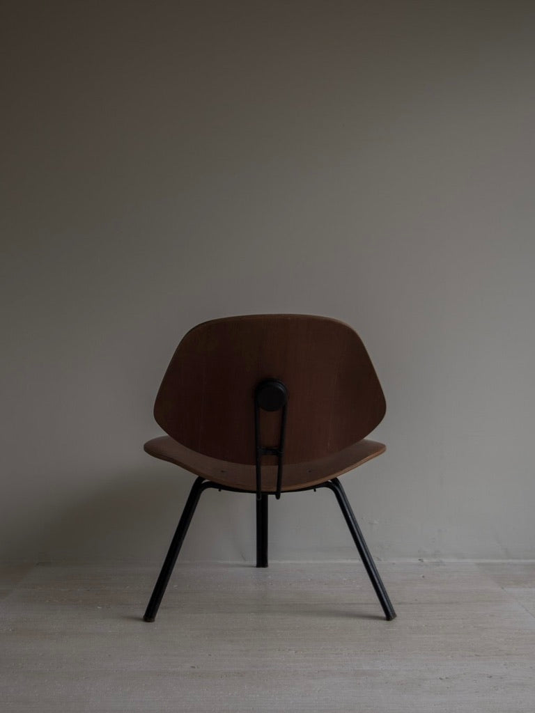 A minimalist wooden chair with black metal legs stands on a light wooden floor against a plain gray wall, evoking an Italian Mid-Century vibe. The room's neutral, modern aesthetic complements the timeless elegance reminiscent of the P31 by Osvaldo Borsani from Bicci de' Medici.