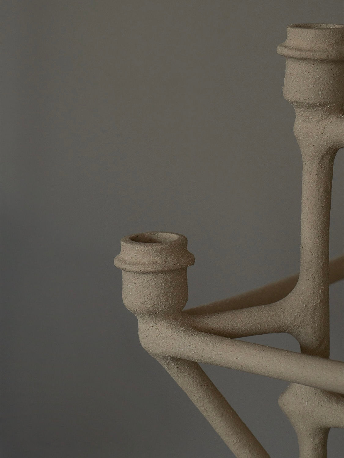 Close-up of the distinctive Clay Chandelier by Solenne Belloir, showcasing its minimalistic design with three holders extending from the center. The textured brown surface stands out against a muted gray-brown background, highlighting the handcrafted details of this unique piece.