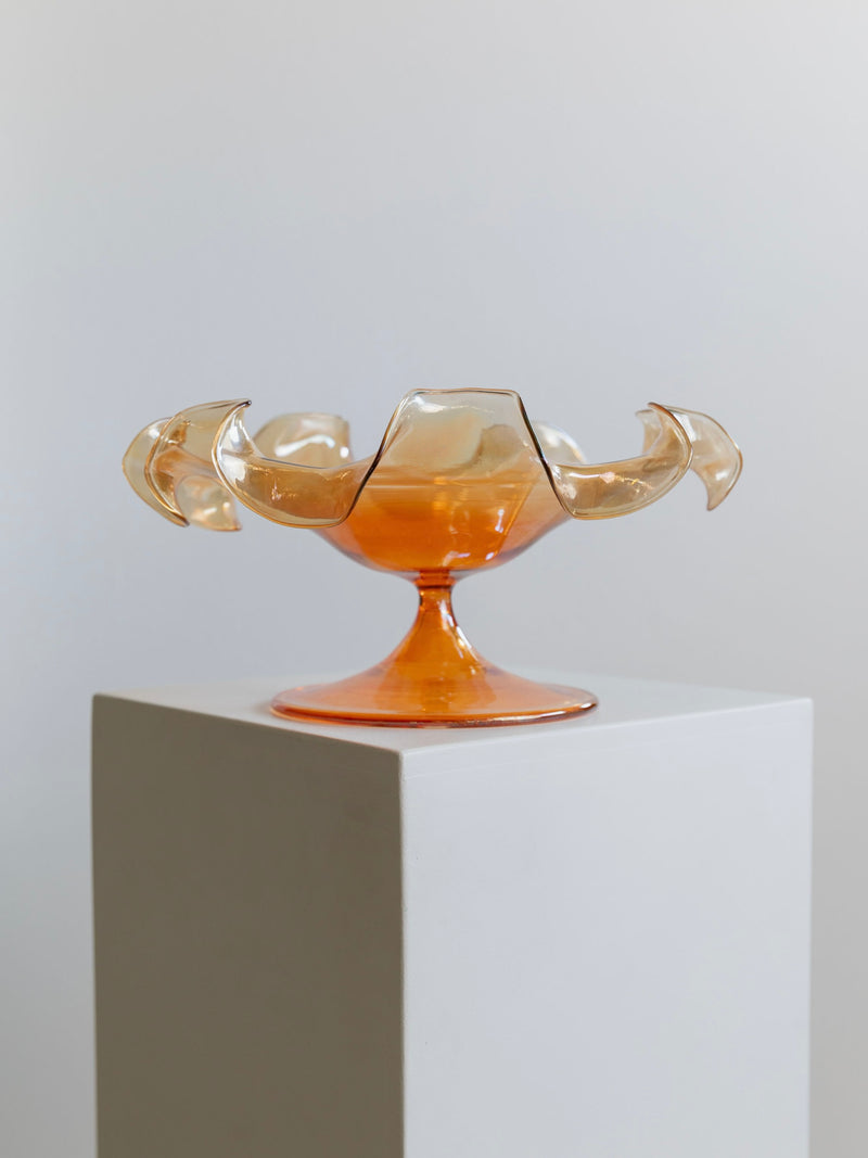 This decorative Murano Glass Bowl by Parise Vetro 70s from Bottega Jacobs boasts an amber hue with artistic, petal-like edges. Accentuated with orange iridescence, it is elegantly showcased on a white pedestal against a neutral background.