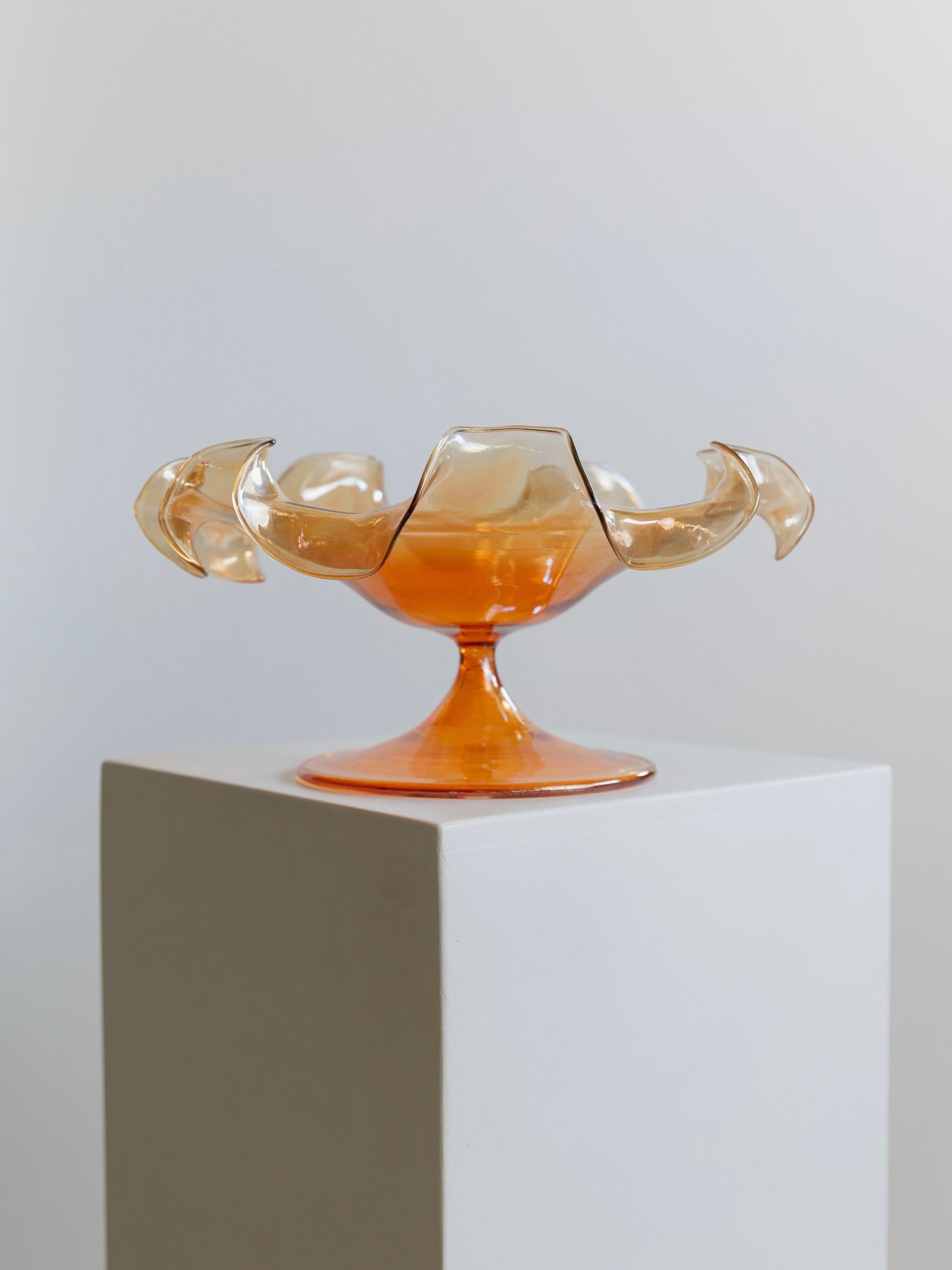 This decorative Murano Glass Bowl by Parise Vetro 70s from Bottega Jacobs boasts an amber hue with artistic, petal-like edges. Accentuated with orange iridescence, it is elegantly showcased on a white pedestal against a neutral background.