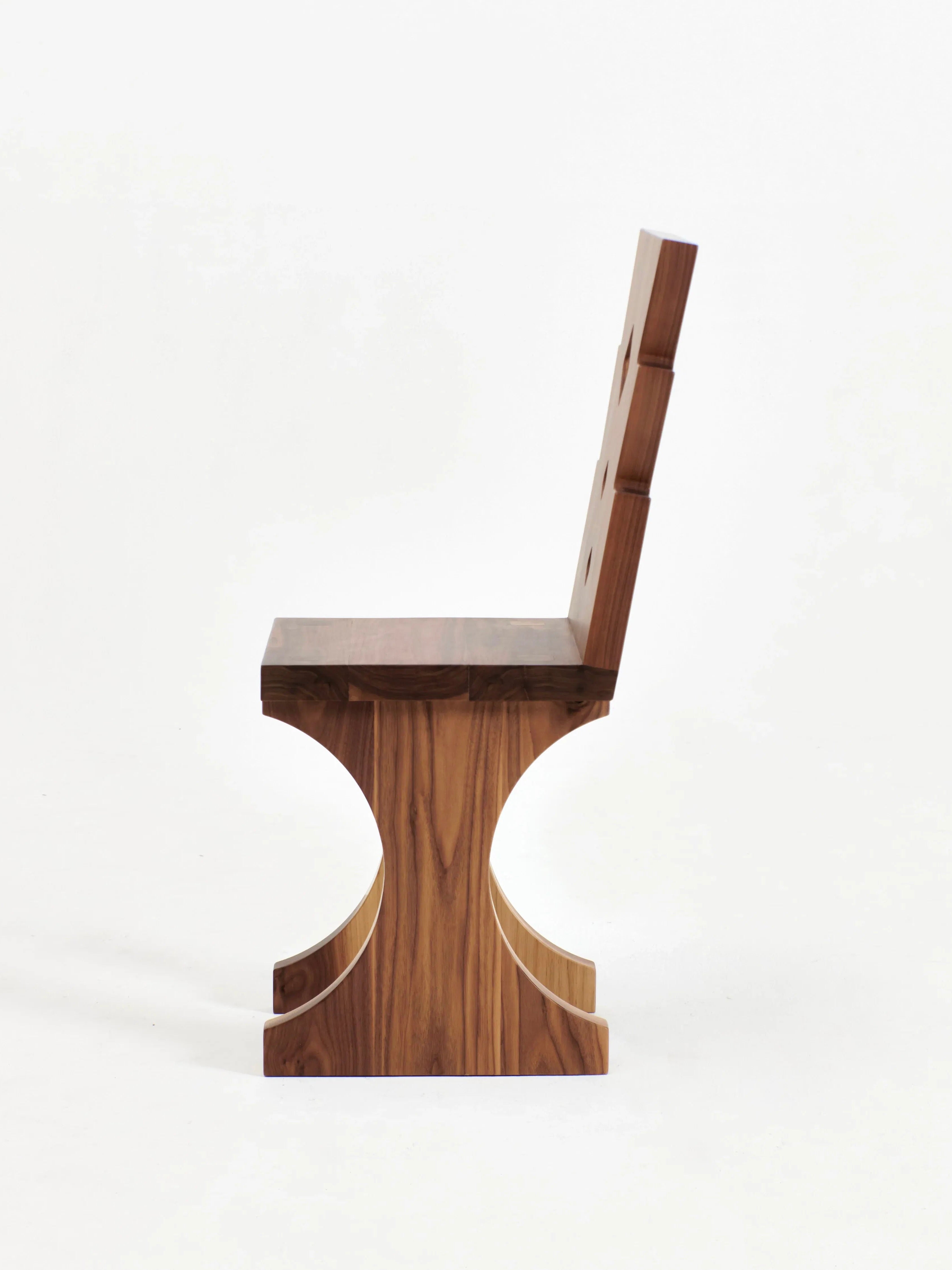 A side view of Project 213A's Portugal Chair No 6 with a modern design, showing a clean, graphic-shaped backrest and a unique base featuring a curved cut-out pattern. Beautifully hand-carved from walnut, the chair showcases a smooth finish and visible natural grain patterns against a plain white background.