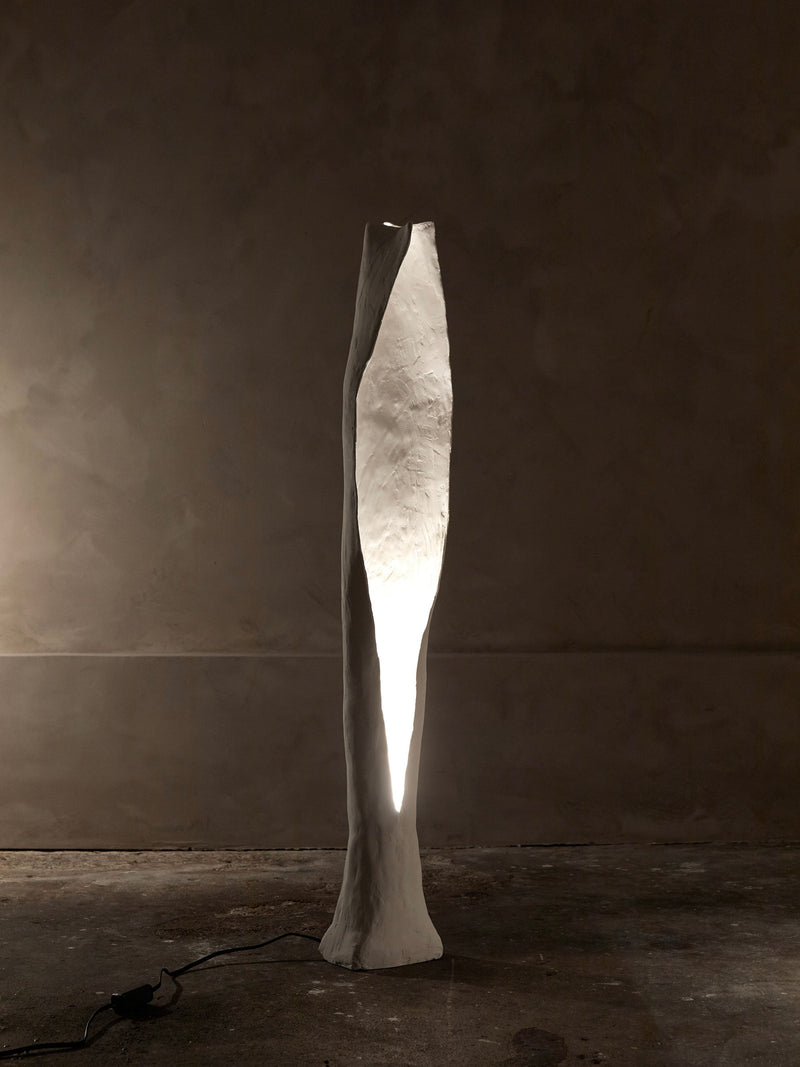 The Alcove Floor Lamp by Studio Tristan Kallas is an elegant, sculptural piece with an organic shape and textured white surface that softly illuminates from its center, standing tall against a dark wall with the visible power cord at its base.