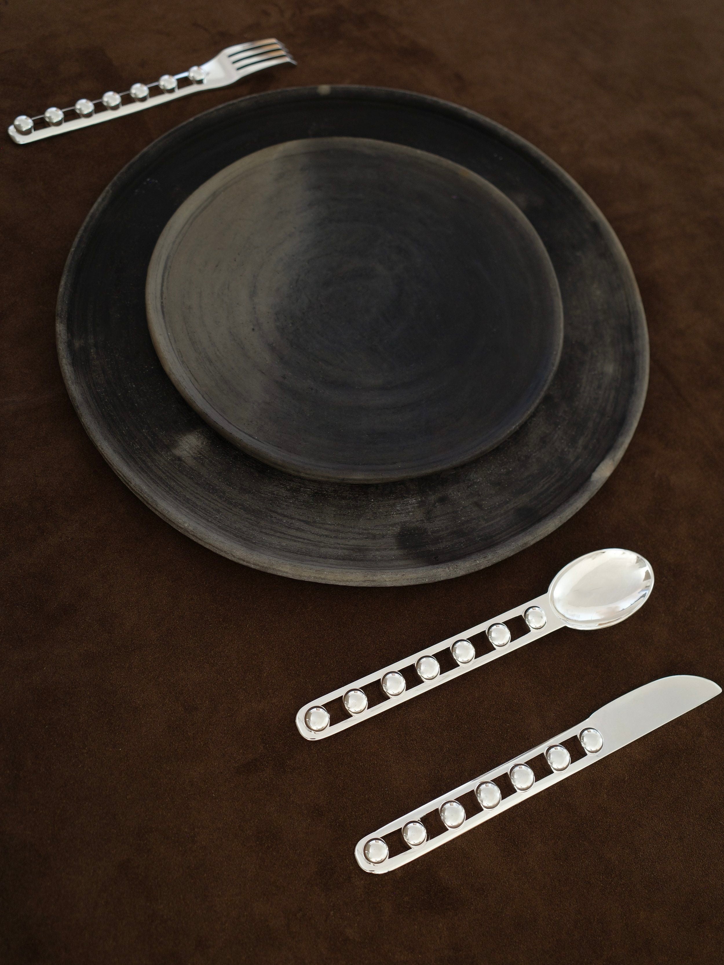 A dark ceramic plate and bowl are arranged on a brown surface, accented by the "Seven Ball" Cutlery Set from Jessi Burch, featuring decorative handles that echo the principles of functional art.