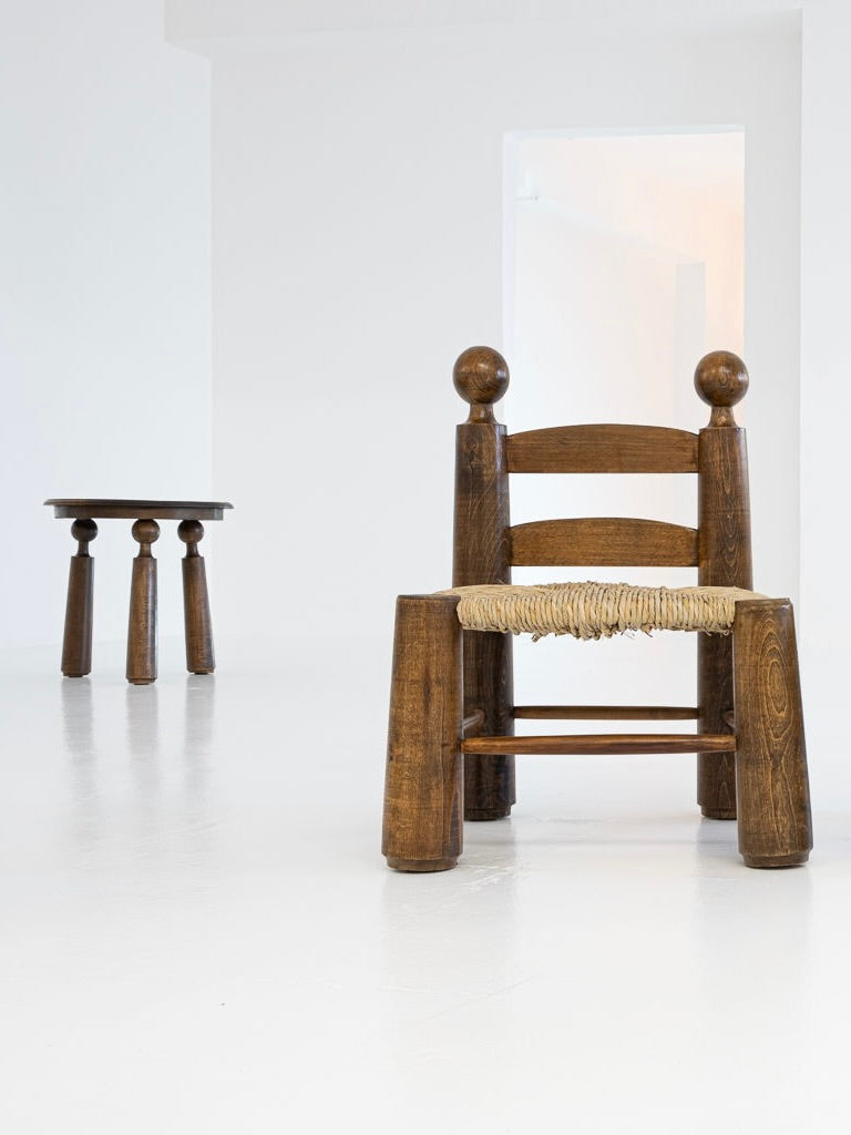 A pair of chairs and a table by Charles Dudouyt, from the 1940s and offered by antibeige, feature in a well-lit, minimalistic room. The wooden chairs have woven seats and rest on a smooth white floor; the sturdy table, with thick rounded legs, highlights Dudouyt's timeless design appeal.