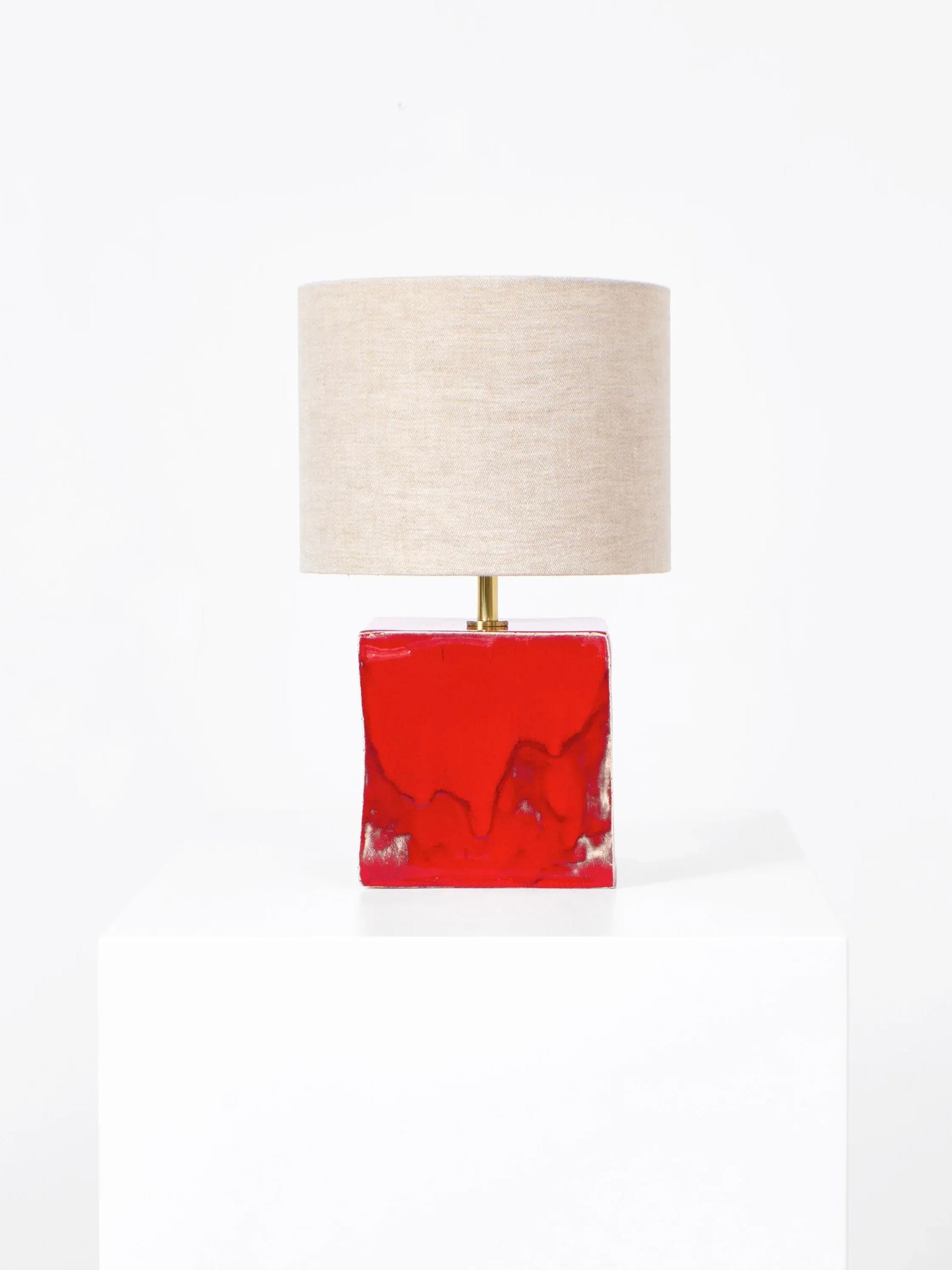 The Arouca Table Light by Project 213A features a beige cylindrical shade on a white pedestal. Its distinctive base, in glossy red ceramic with an artistic shape, offers bespoke options. Displayed against a minimalist white background for elegant presentation.