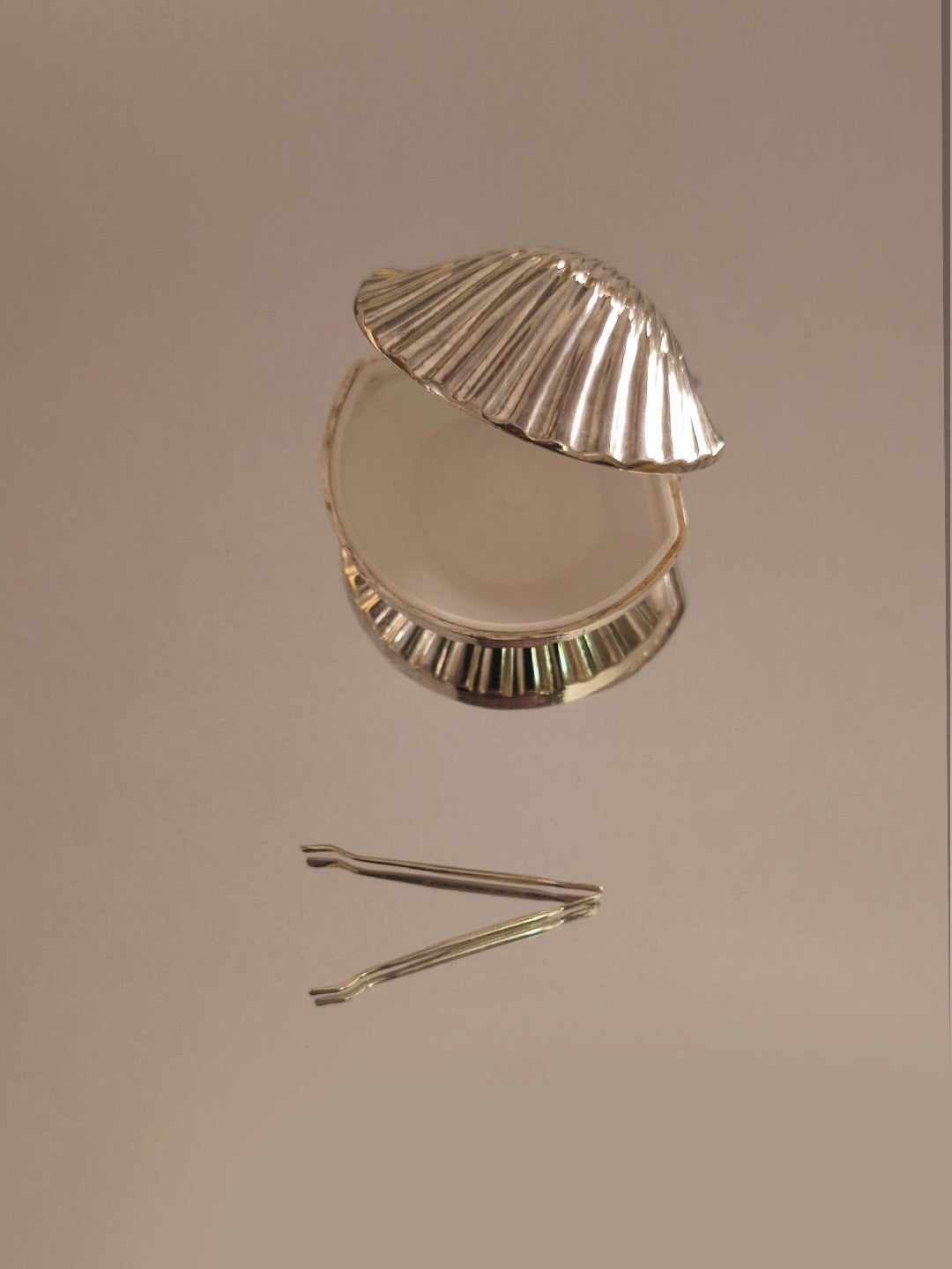 The Small Shell Box by Les Objoies, with its silver-plated design, lies open on a beige surface, resembling a delicate jewelry storage piece, accompanied by metallic tweezers positioned below.