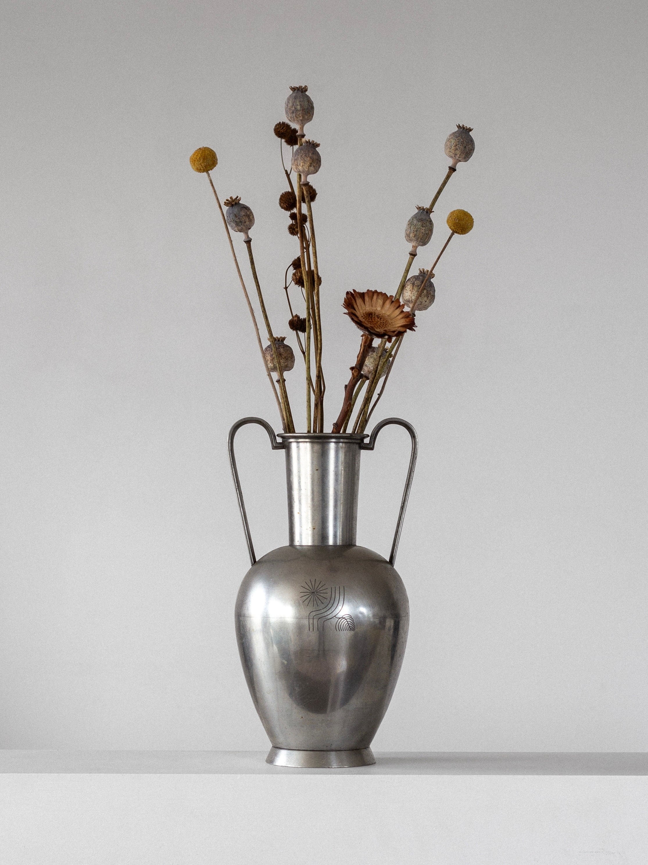 The Large Modernist Metal Amphora by Spigel, a 1930s Scandinavian piece, displays dried flower arrangements with yellow billy balls and poppy pods in a silver vase against a plain gray background on a white surface.