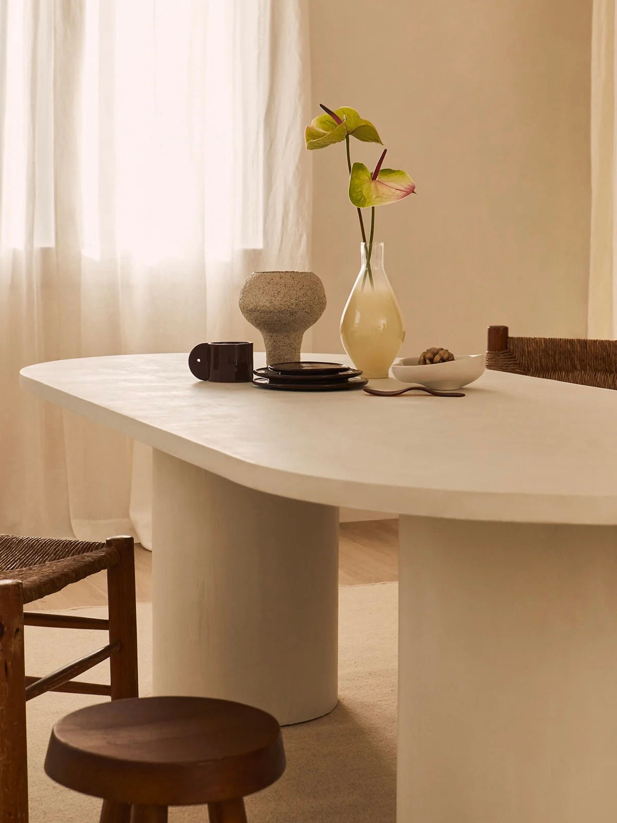 A minimalist dining area features the MarlotBaus Reye Dining Table, a handcrafted oval microcement piece. Adorning it are a vase with green and pink flowers, a fruit bowl, and a textured decor item. Sheer curtains gently diffuse natural light throughout the space.