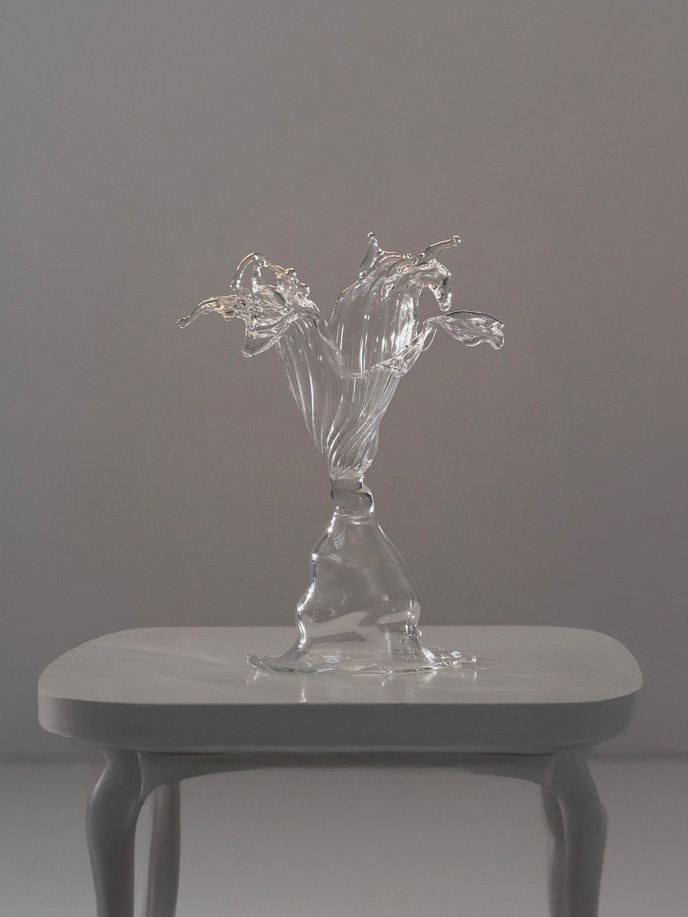 The "Objects of Affection_Romance" glass sculpture by Pia Glassworks, with its flowing, twisted form on a simple wooden table, catches the light beautifully. Its translucent curves reflect subtly against the plain gray background, highlighting its elegance as a talisman of imagination.