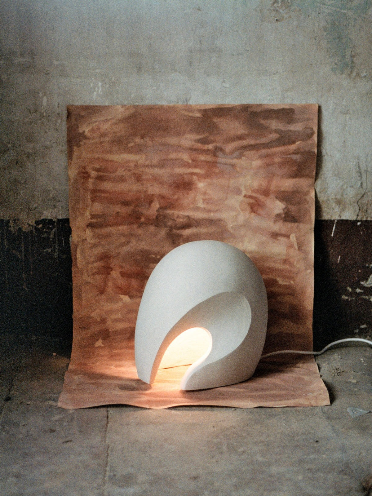 The Coquille Ceramic Lamp by Lea Bigot features a unique shape that casts a warm glow against an earthy brown background. This handmade piece has a rustic, unfinished look with part of the power cord visible on the floor.