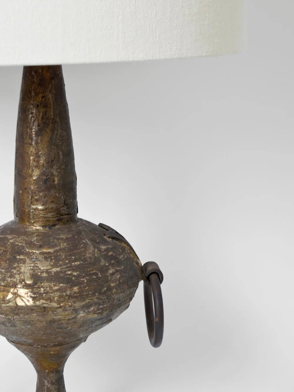 A detailed image showcases the textured base of the Max Bre Gilded Iron Brutalist Table Lamp by Barracuda Interiors, designed in the 1960s, featuring a distinctive curved metal ring. The lamp is complemented by a neutral lampshade peeking from the top, all set against an understated gray backdrop.