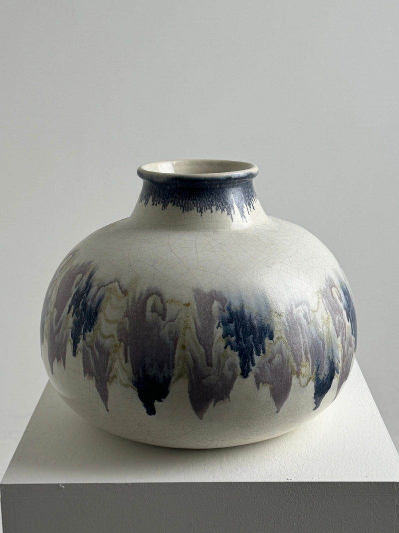 On a white pedestal sits a large ceramic vase by Figuresdesign, crafted by Ateliers Primavera. It boasts a round shape with a narrow opening and features a cracked glaze adorned with blue and gray wavy patterns against a light backdrop. The minimalistic setting is enhanced by soft lighting.