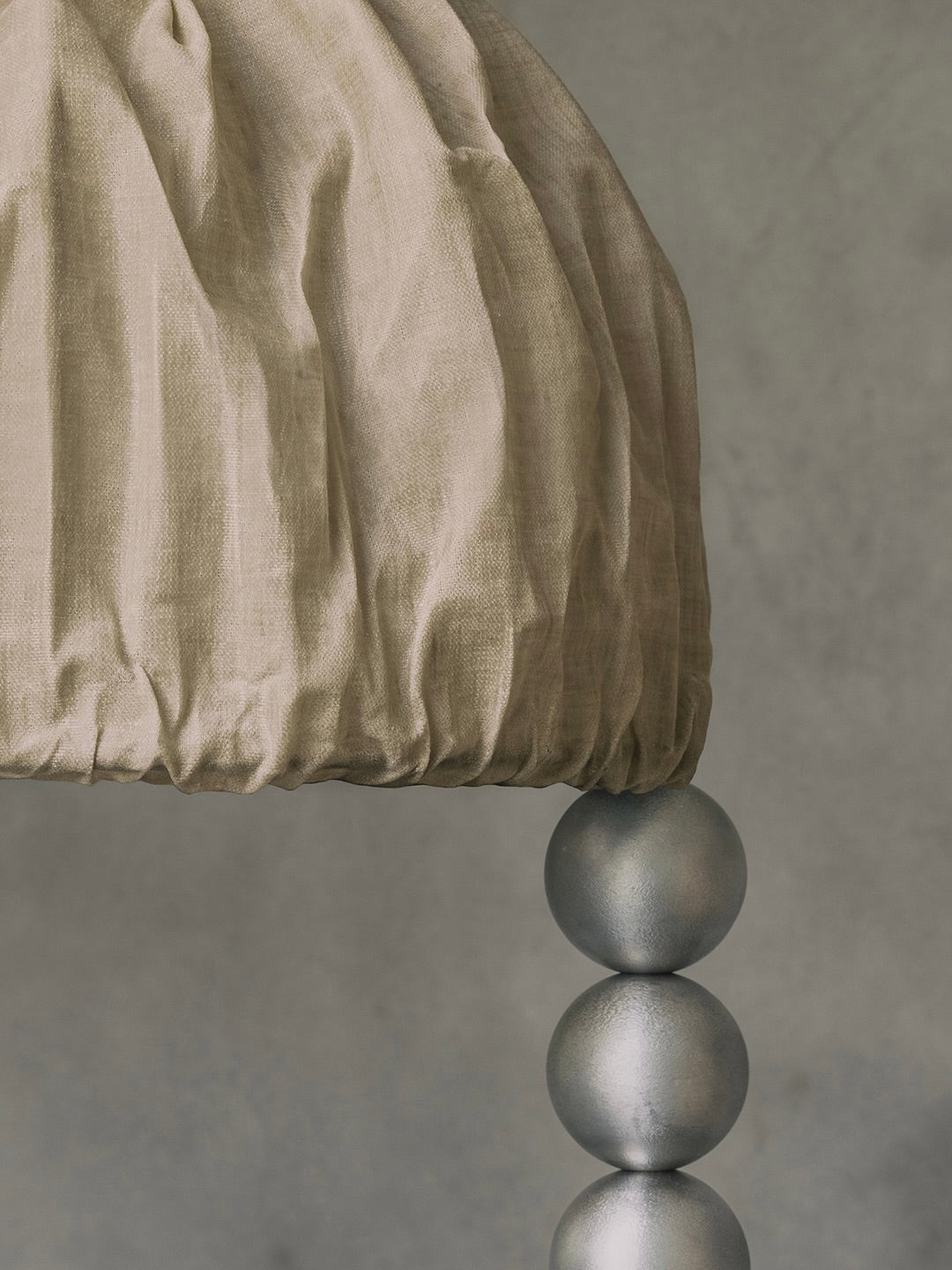 A close-up of the Kaytar "ALTER" Floor Light showcases a textured beige fabric dome atop three stacked sand-cast aluminum spheres, set against a soft, muted gray background.