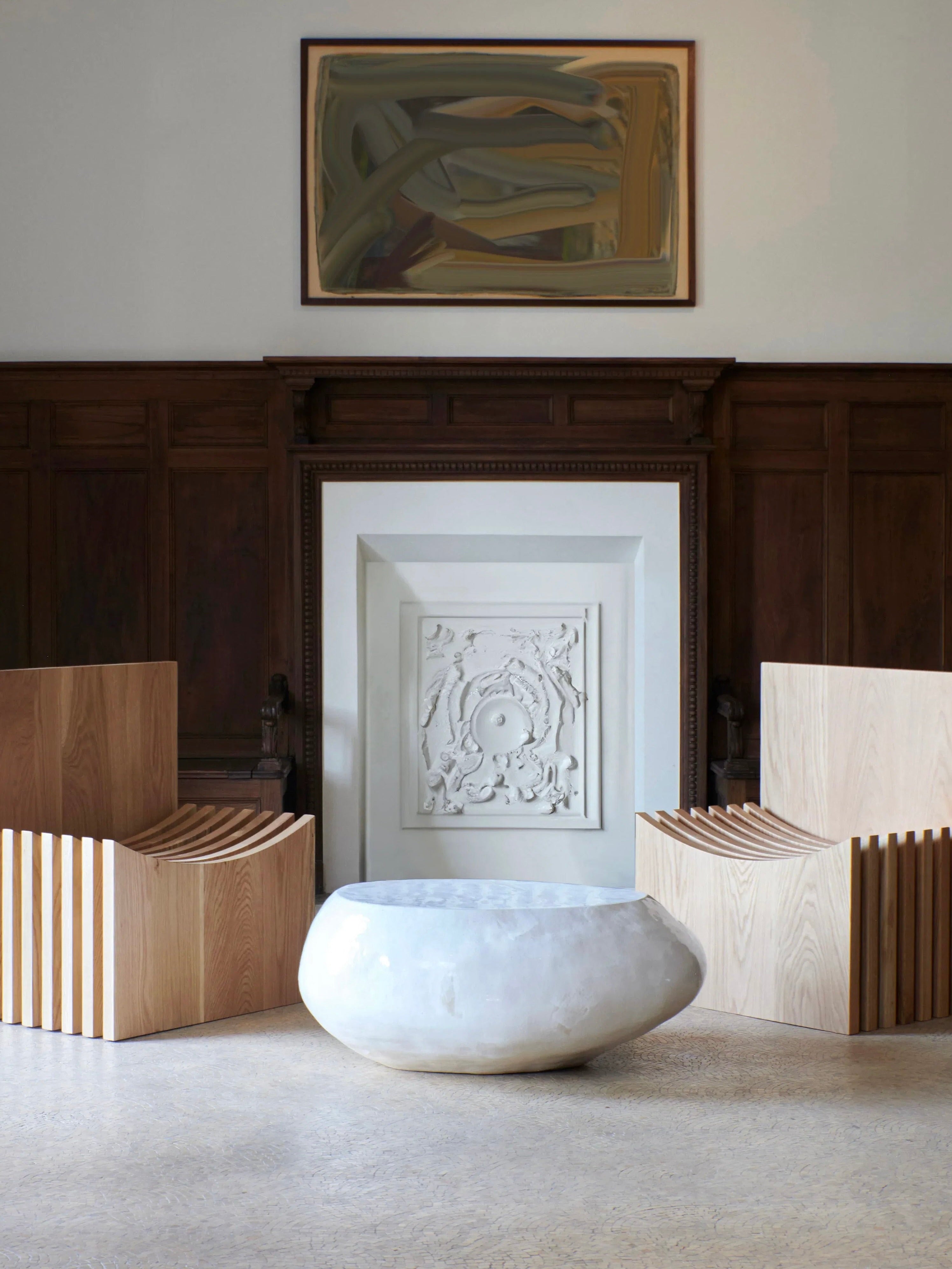 A minimalist room features two wooden chairs with slatted backs, a round Ceramic Coffee Table in Cream by Project 213A with customized glaze, and a large decorative fireplace with intricate molding. Above the fireplace hangs an abstract framed painting. The walls are partially lined with dark wood paneling.