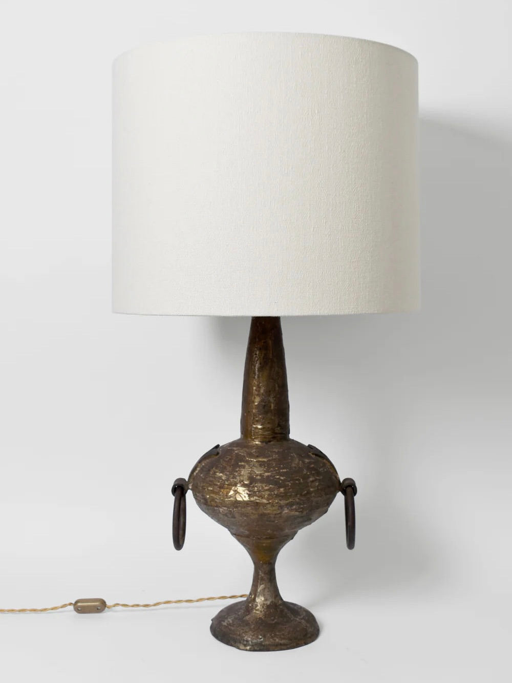 The Max Bre Gilded Iron Brutalist Table Lamp by Barracuda Interiors from the 1960s features a large white fabric shade paired with a textured, metallic base resembling an ancient vase, and adorned with two circular metal handles. This neoclassical lamp includes a twisted cord and presents itself elegantly on a white background.