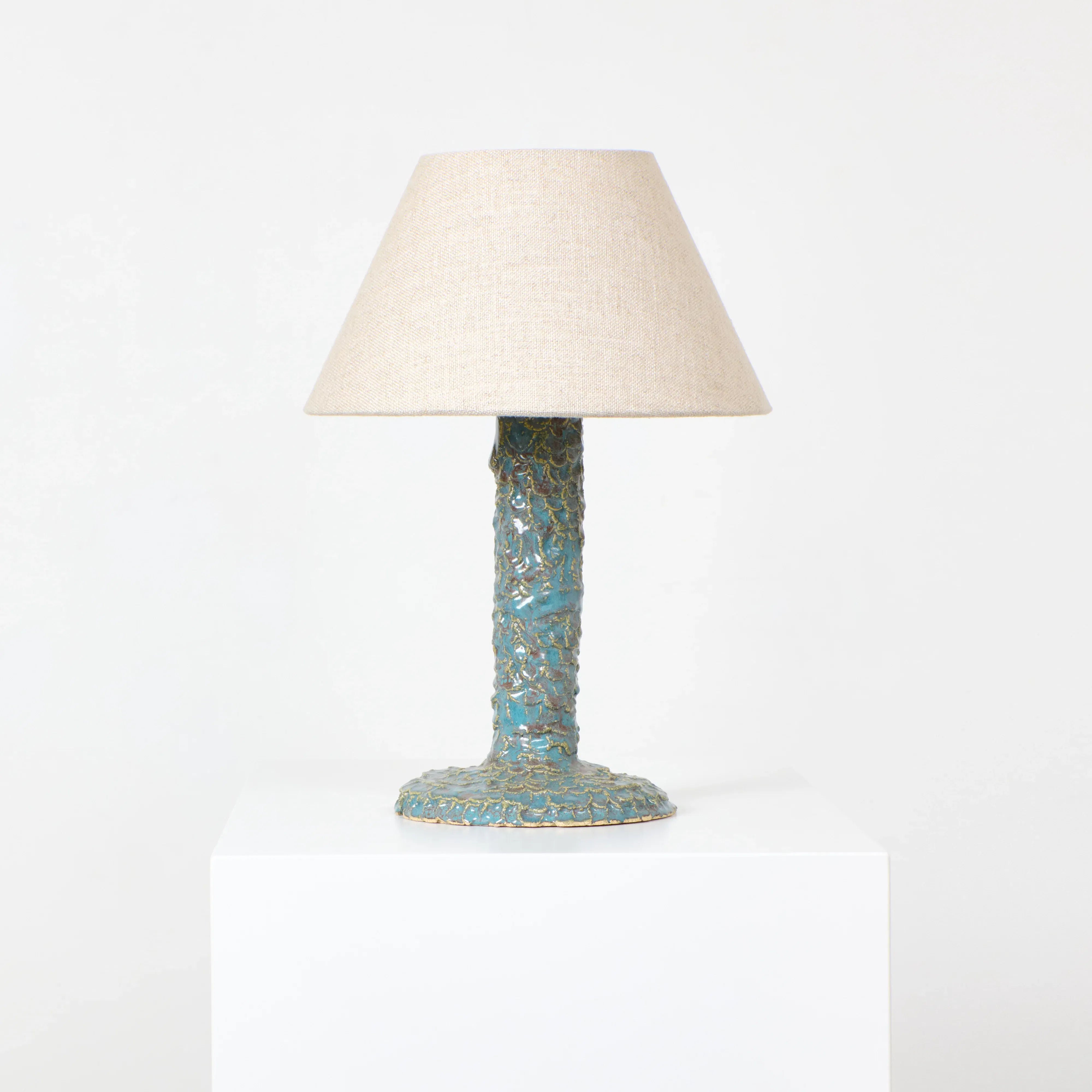 Project 213A's Casa Bedside Light, featuring a textured blue-green ceramic base and a beige conical fabric lampshade, stands elegantly on a white pedestal against a plain white backdrop.