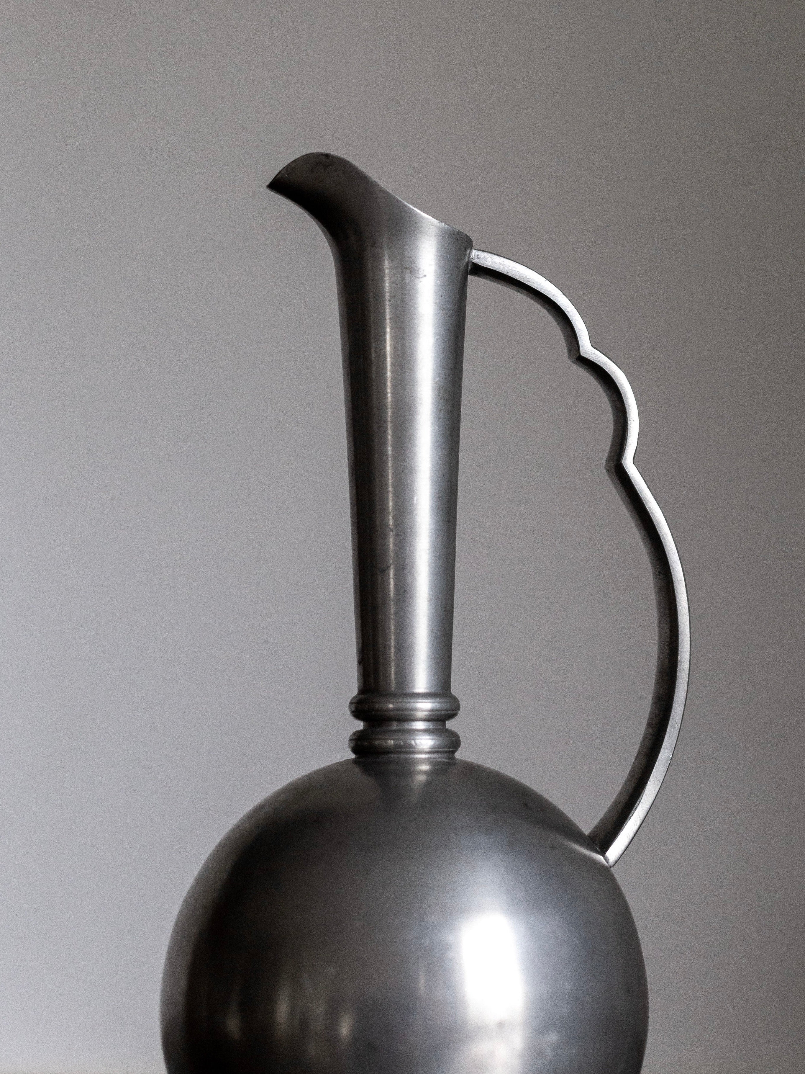 A Spigel Art Deco Metal Vase from the 1930s, featuring a tall, slender neck and curved handle, is depicted against a plain background. The rounded bottom contrasts with the sleek, tapered top, lending a blend of Swedish Art Deco aesthetic to the vessel.