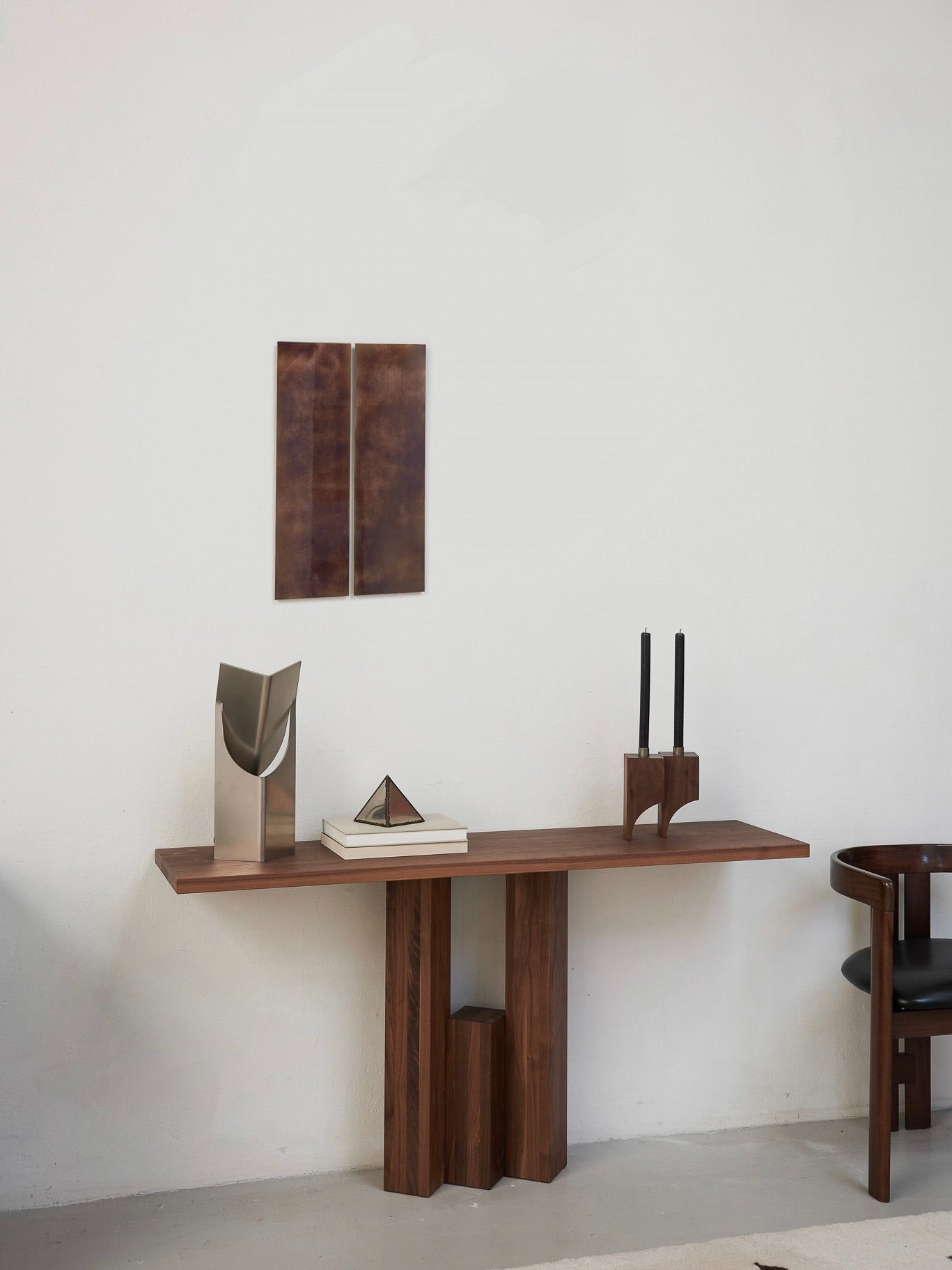 A minimalist room showcases a Mokko Fenestra Console against a white wall. Atop are a metal sculpture, books, geometric object, and candle holder. Above is abstract brown wall art while a dark wooden chair on the right adds customizable design flair.