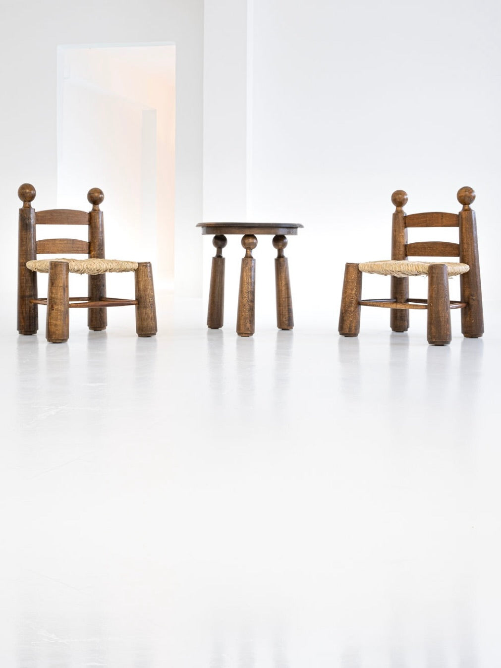 A glossy white floor features a minimalist, brightly lit setup with antibeige's 1940s Charles Dudouyt Set of 2 Chairs and 1 Table. The chairs, with rounded backrests and cushioned seats, complement the solid oak table with stubby legs.