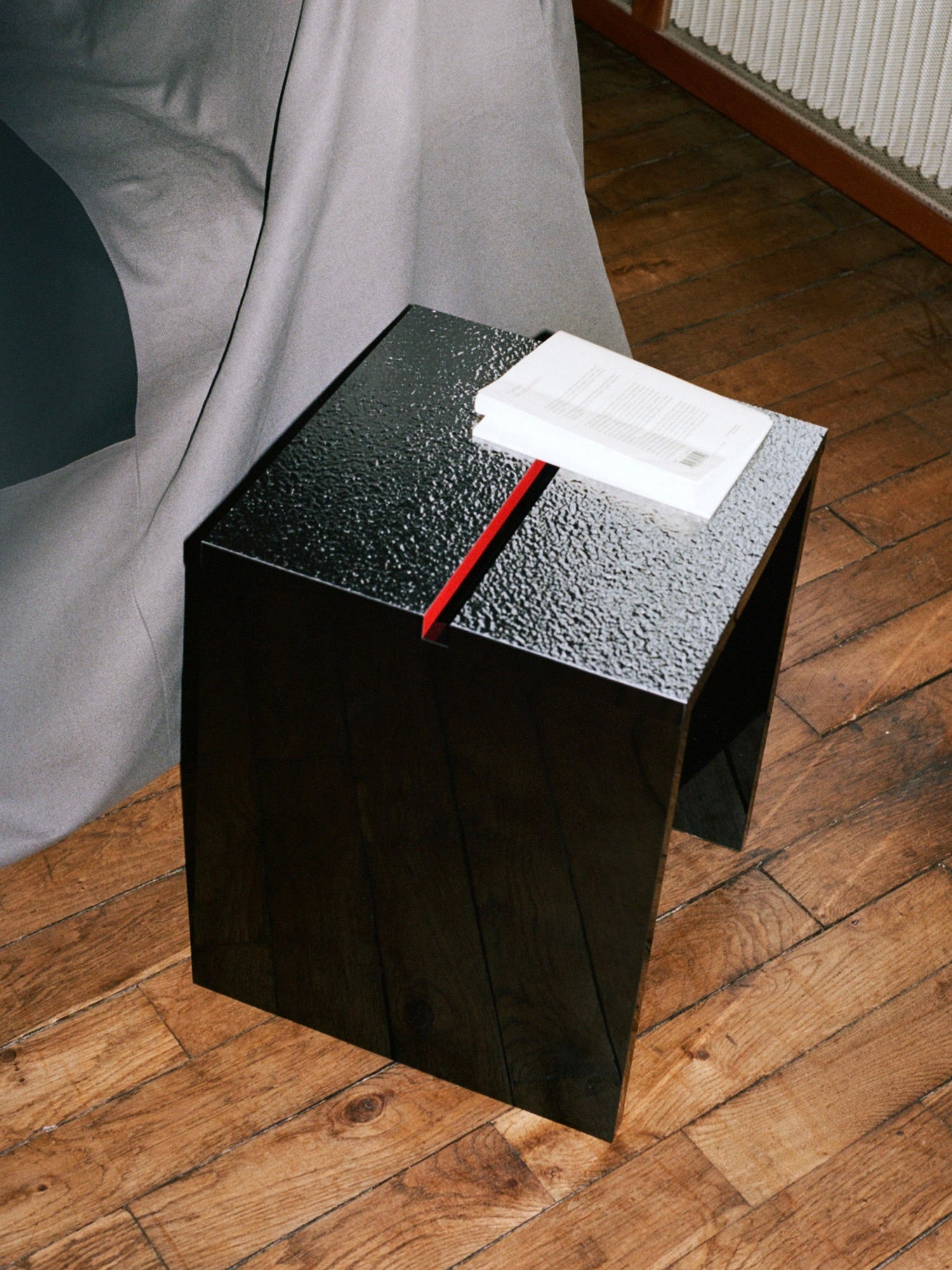 A sleek, glossy black Mara Gap Stool – Wet Black by TEGET sits on a wooden floor with a red accent line. A white book with text rests on top. In the background, draped gray fabric and part of a radiator complete this scene of customizable furniture elegance.