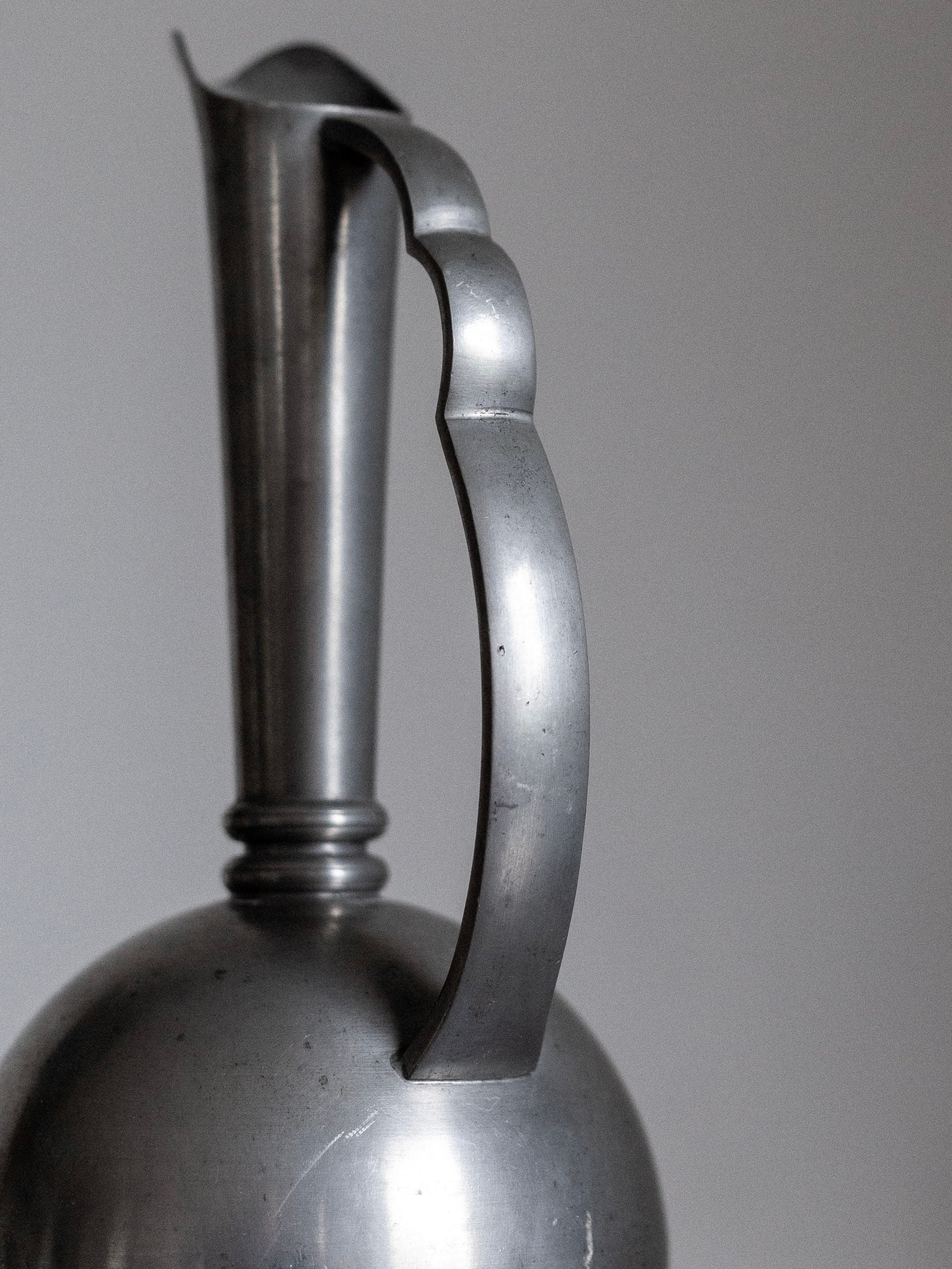 The image displays a close-up of the Art Deco Metal Vase from the 1930s by Spigel. This collectible pewter piece, evoking the Swedish Art Deco style of CG Hallberg, features a smooth, polished surface and a tall, narrow spout with a gracefully curved handle. The plain gray background accentuates the simplicity and elegance of its design.